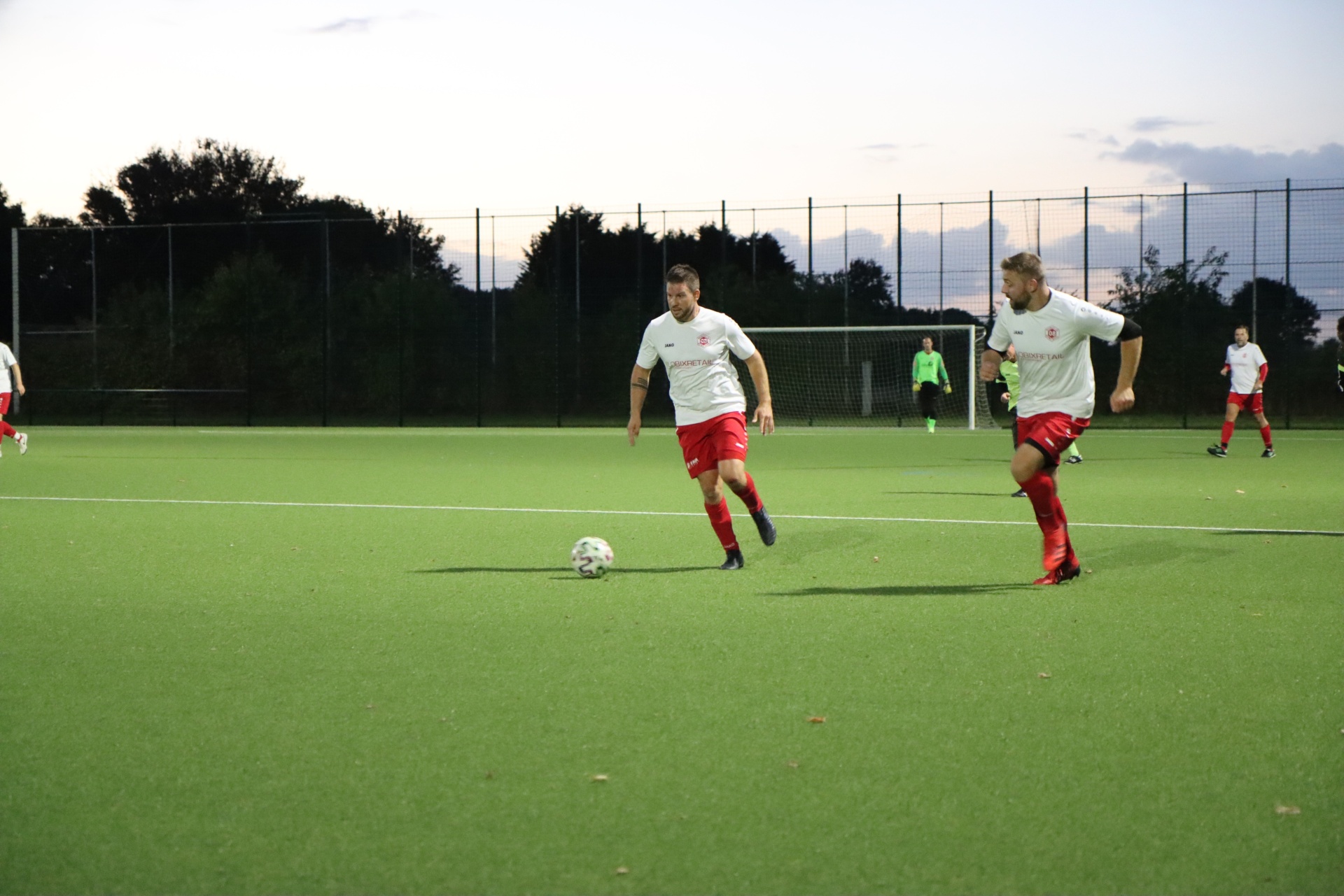[{"clang_id":1,"value":"Fußballfeld mit zwei Spielern in weißen und roten Trikots, die einen Ball spielen. Im Hintergrund weitere Spieler und ein Zaun."}]