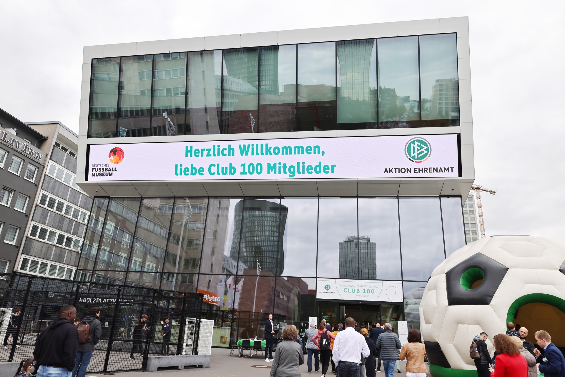 [{"clang_id":1,"value":"Gebäudefront mit großem Banner \"Herzlich Willkommen, Liebe Club 100 Mitglieder\". Menschengruppe davor, teilweise vor überdimensionalem Fußball."}]