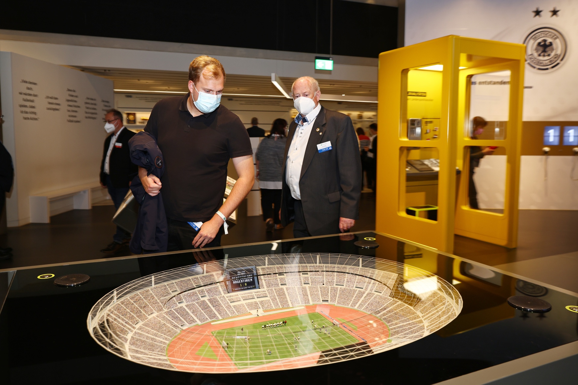[{"clang_id":1,"value":"Zwei Männer betrachten ein Modell eines Fußballstadions in einem Museum. Beide tragen Masken. Im Hintergrund weitere Besucher und Ausstellungsstücke."}]