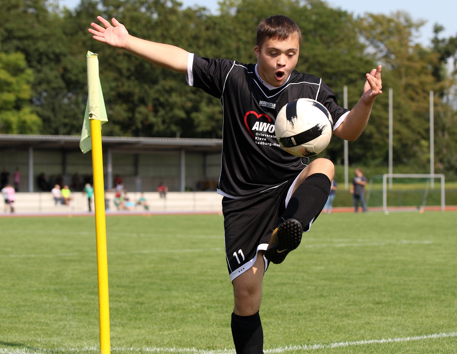 [{"clang_id":1,"value":"Fußballspieler in schwarzem Trikot und Shorts schießt einen Fußball auf einem grünen Rasenfeld. Im Hintergrund ein Wald und weitere Personen."}]