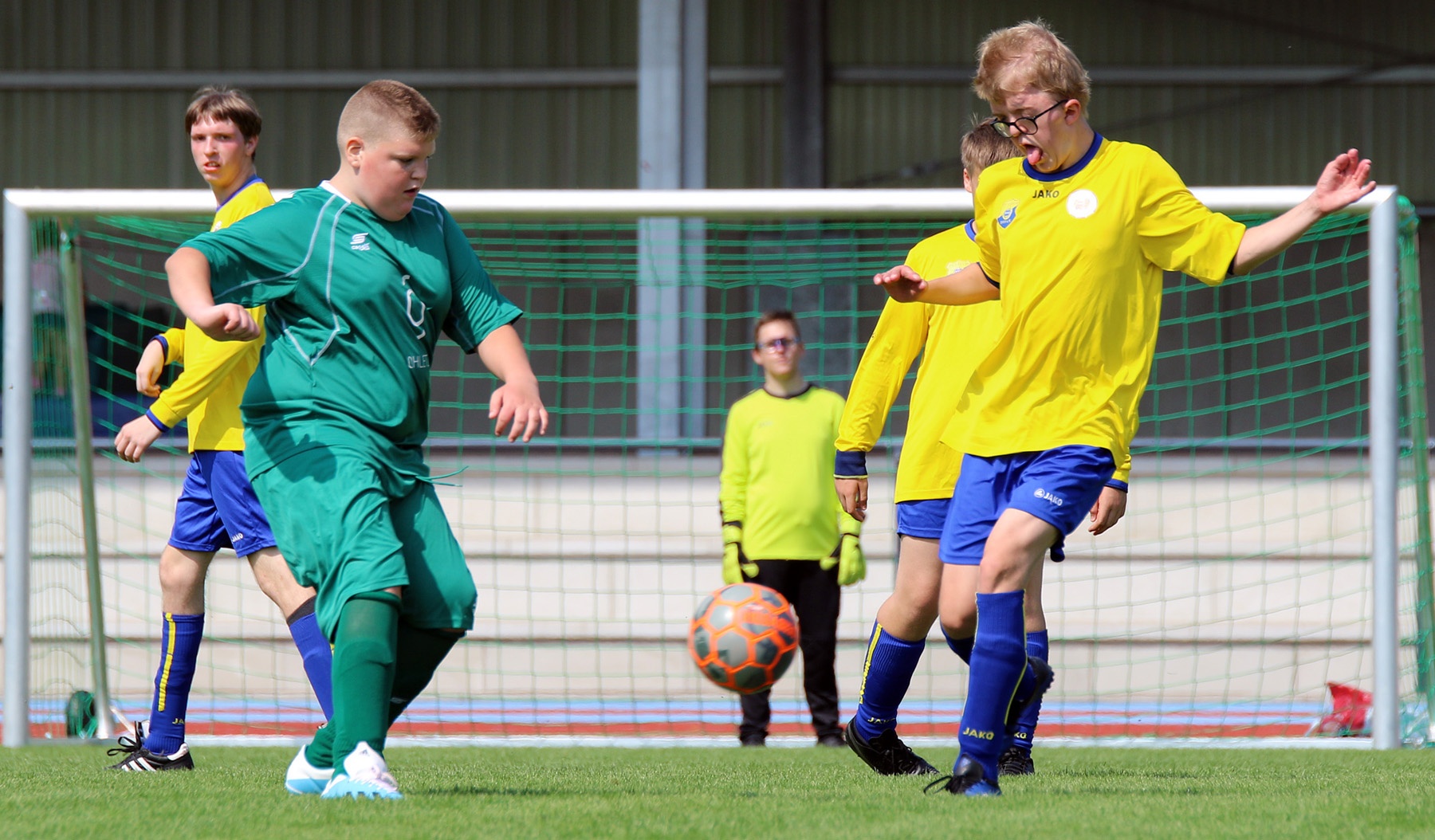 [{"clang_id":1,"value":"Fußballspiel: Zwei Spieler kämpfen um den Ball vor dem Tor. Im Hintergrund weitere Spieler und ein metallenes Vereinsheim."}]