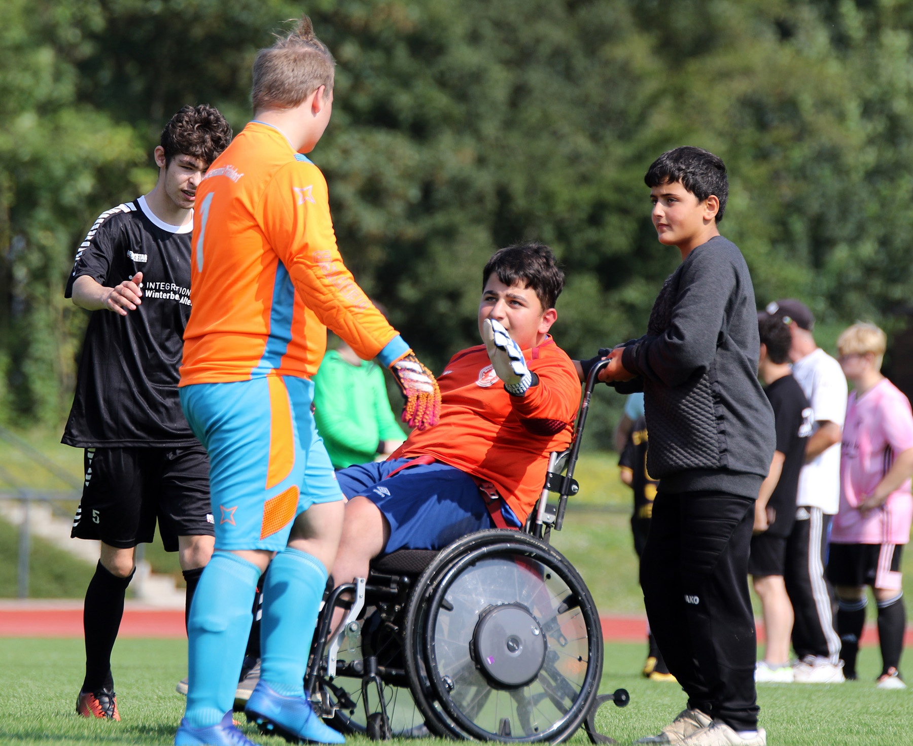 [{"clang_id":1,"value":"Fußballfeld: Vier Jugendliche um einen Rollstuhlfahrer. Zwei in Fußballkleidung reichen ihm die Hände, ein weiterer steht daneben. Im Hintergrund weitere Personen."}]