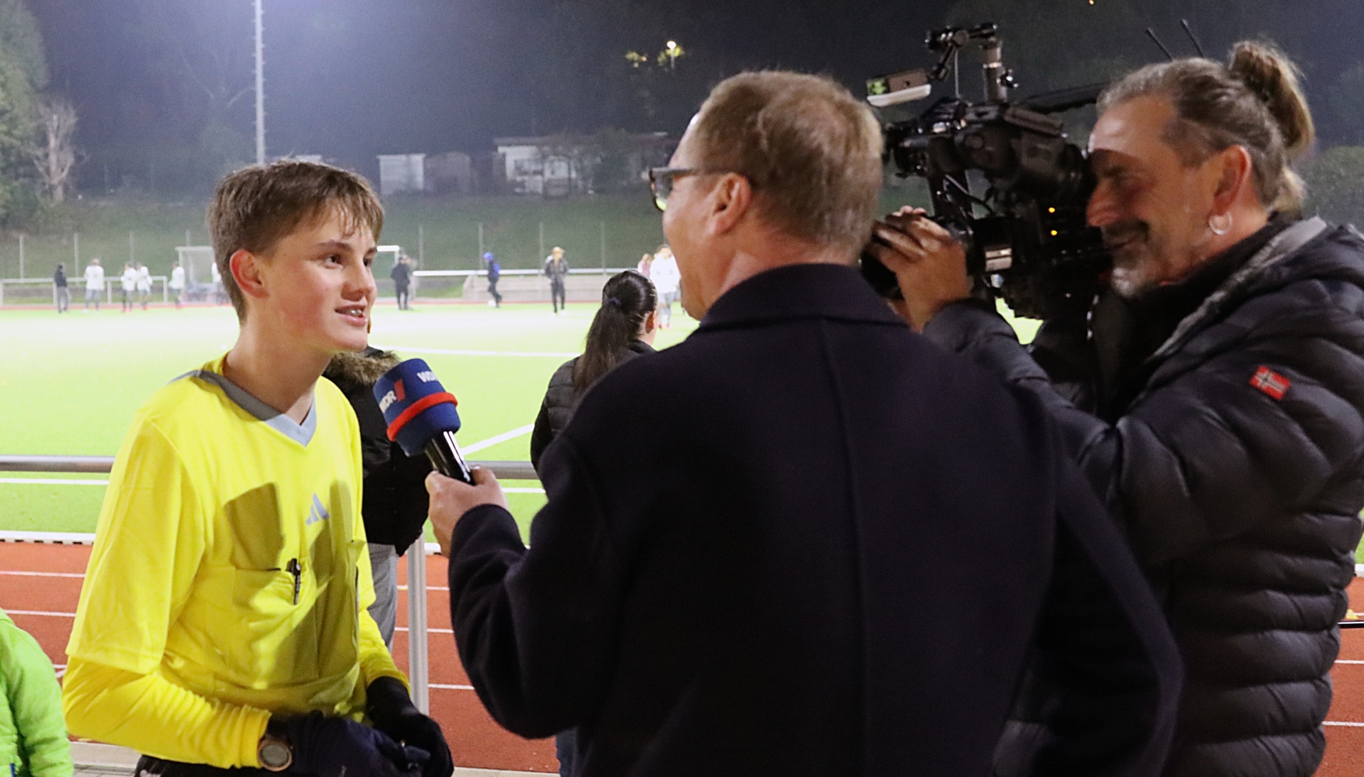 [{"clang_id":1,"value":"Junge im Fußballtrikot wird interviewt. Interviewer hält Mikrofon. Kamerateam filmt im Hintergrund auf einem Sportplatz bei Nacht."}]