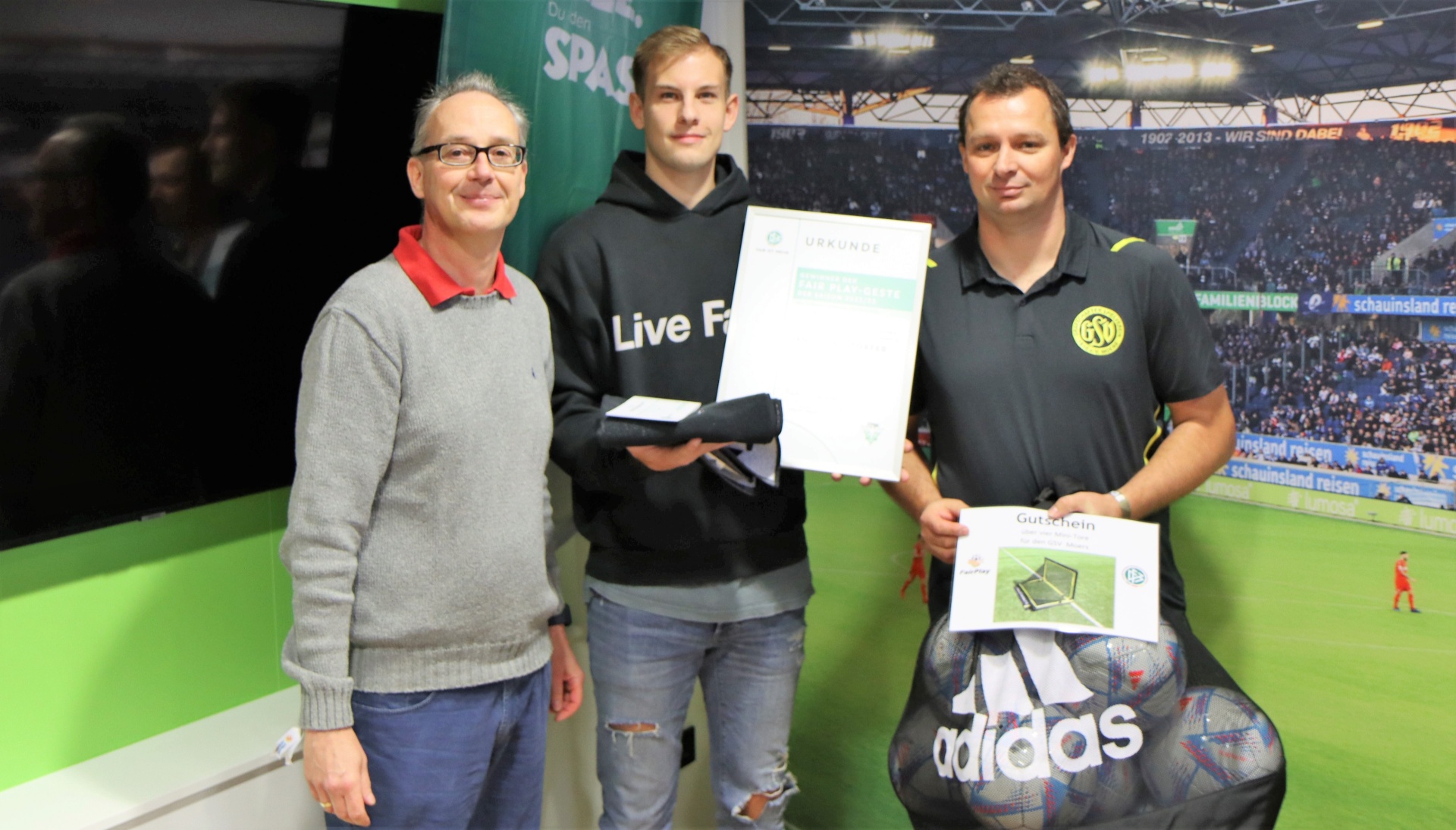 [{"clang_id":1,"value":"Drei Männer stehen nebeneinander und halten Urkunden. Im Hintergrund eine Indoor-Fußballhalle mit Zuschauerrängen."}]