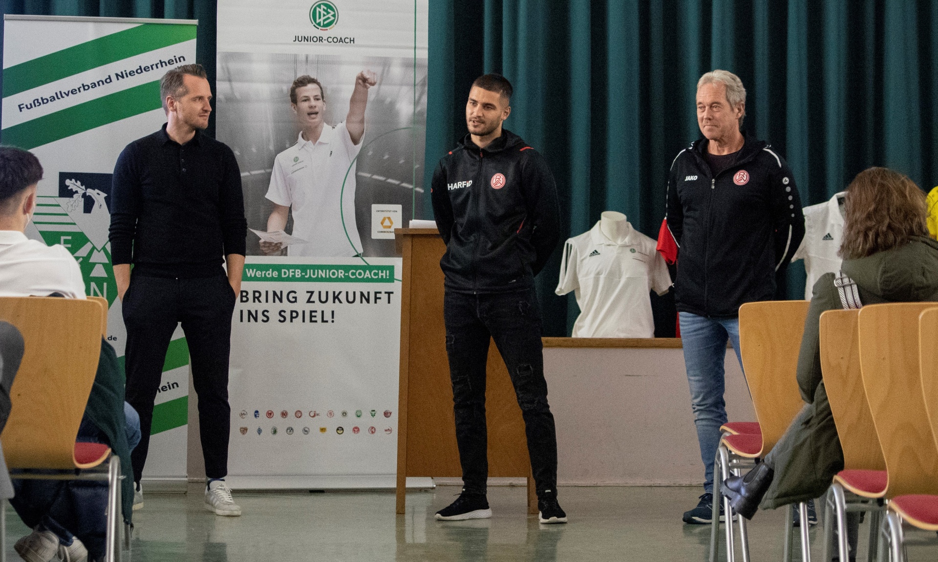 [{"clang_id":1,"value":"Drei Männer stehen in einem Raum vor einem Banner mit Fußballmotiven. Links und rechts sitzen Personen. Einer der Männer spricht ins Publikum."}]