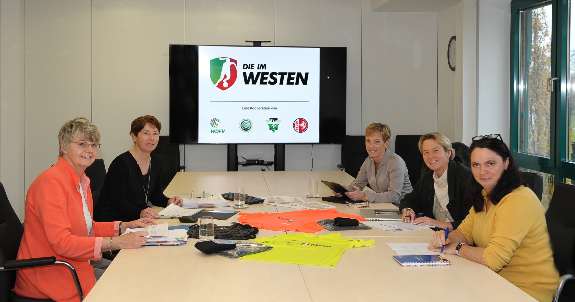 [{"clang_id":1,"value":"Sitzung in einem Konferenzraum: Sechs Frauen sitzen an einem großen Tisch. Im Hintergrund eine Präsentation mit dem Titel \"DIE IM WESTEN\"."}]