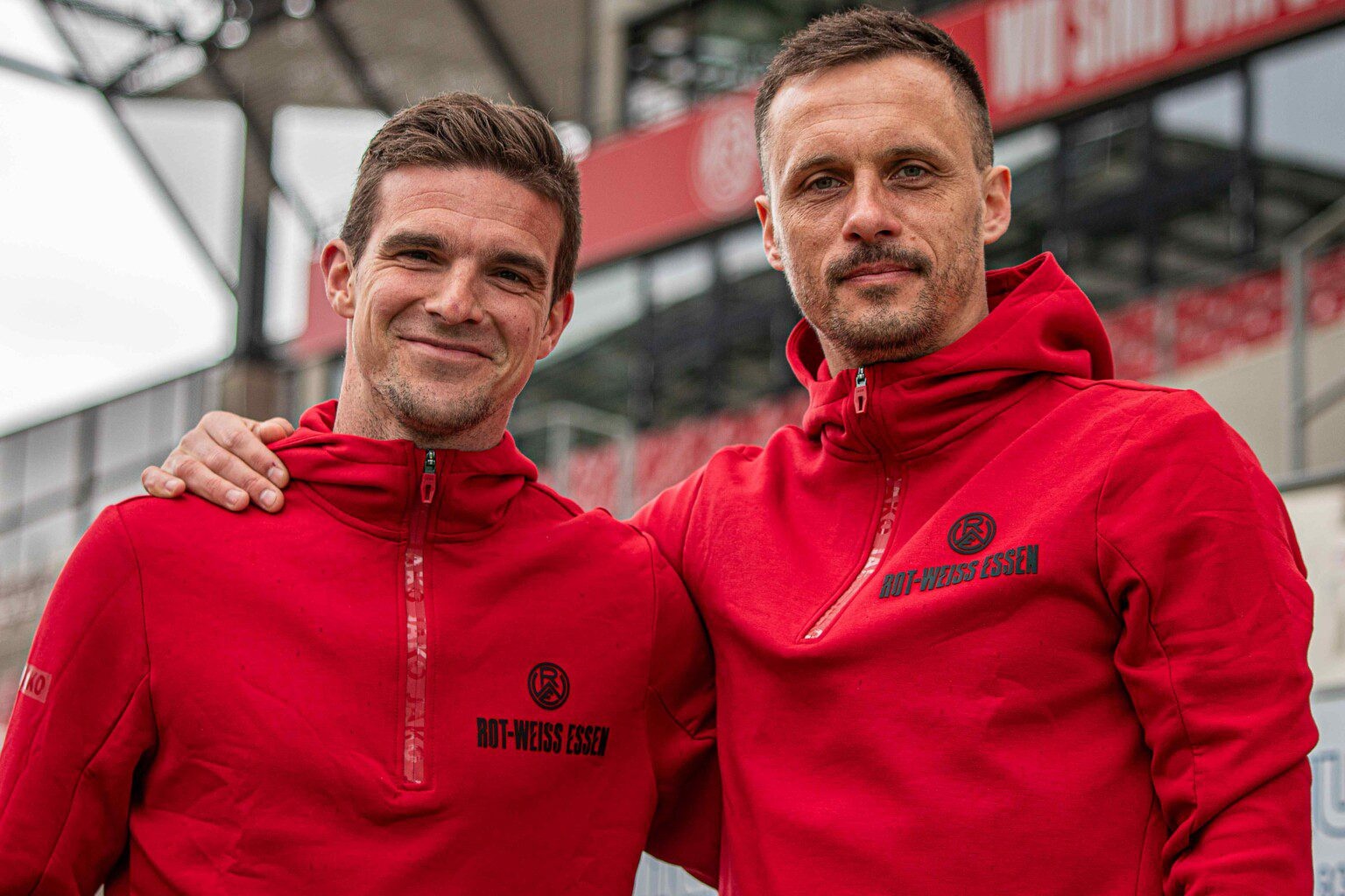 [{"clang_id":1,"value":"Zwei Männer mit kurzgeschorenen Haaren, beide mit Trainingsanzügen von Rot-Weiss Essen, stehen lächelnd vor einem Fußballstadion."}]