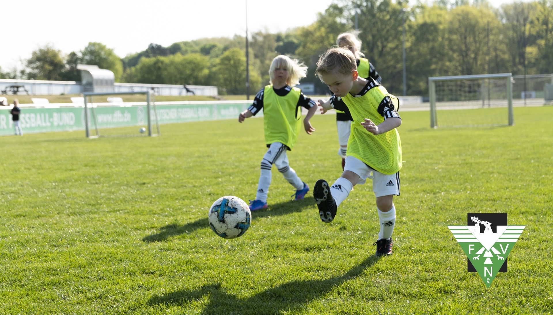 [{"clang_id":1,"value":"Junge Fussballspieler mit gelben Westen trainieren auf grünem Rasenfeld. Ein Kind dribbelt den Ball. Im Hintergrund Begrenzung und Zuschauertribüne."}]