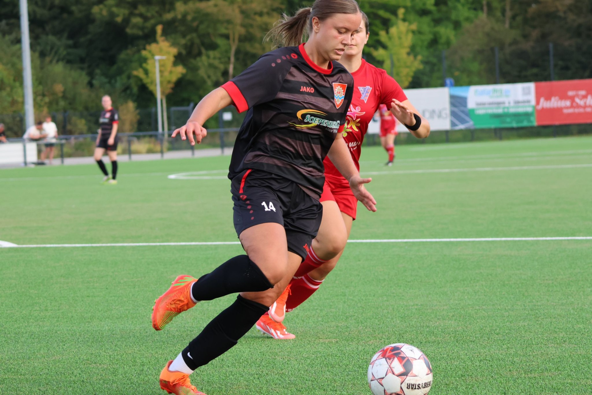 [{"clang_id":1,"value":"Fußballspielerin mit Nummer 14 in schwarzem Trikot dribbelt den Ball auf grünem Rasen. Im Hintergrund weitere Spieler und Zuschauer."}]