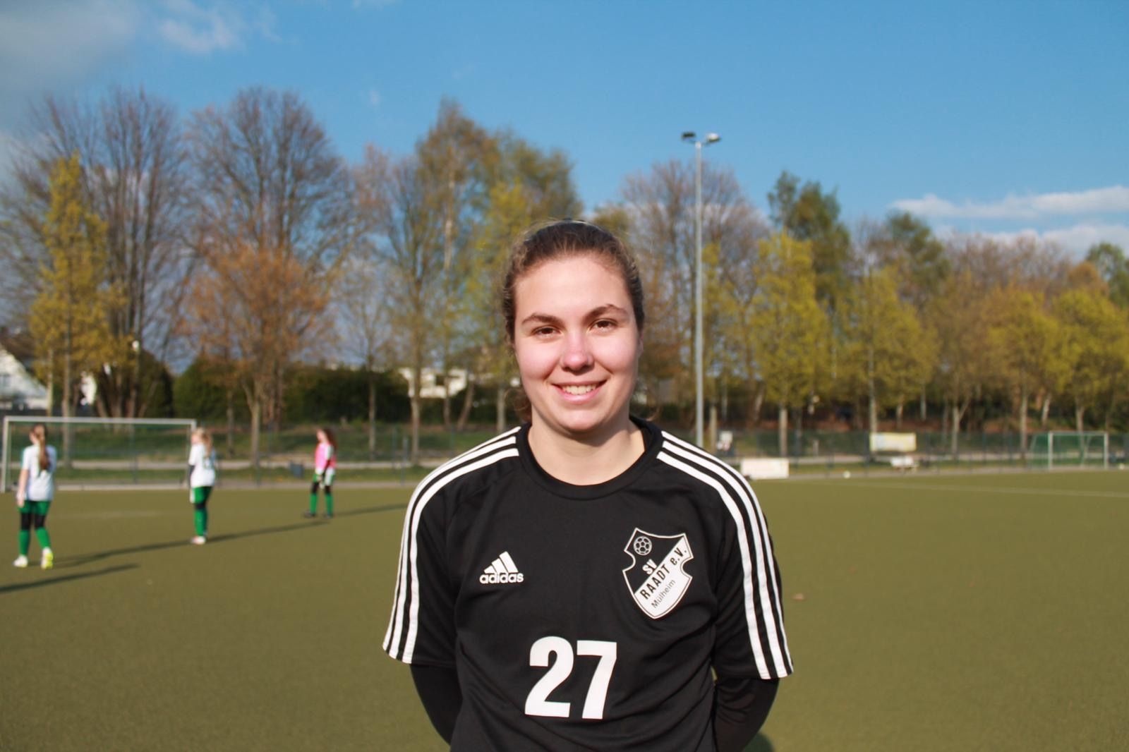 [{"clang_id":1,"value":"Fußballspielerin mit Nummer 27 auf schwarzem Trikot, lächelnd auf einem Fußballfeld. Im Hintergrund Bäume und andere Spieler."}]