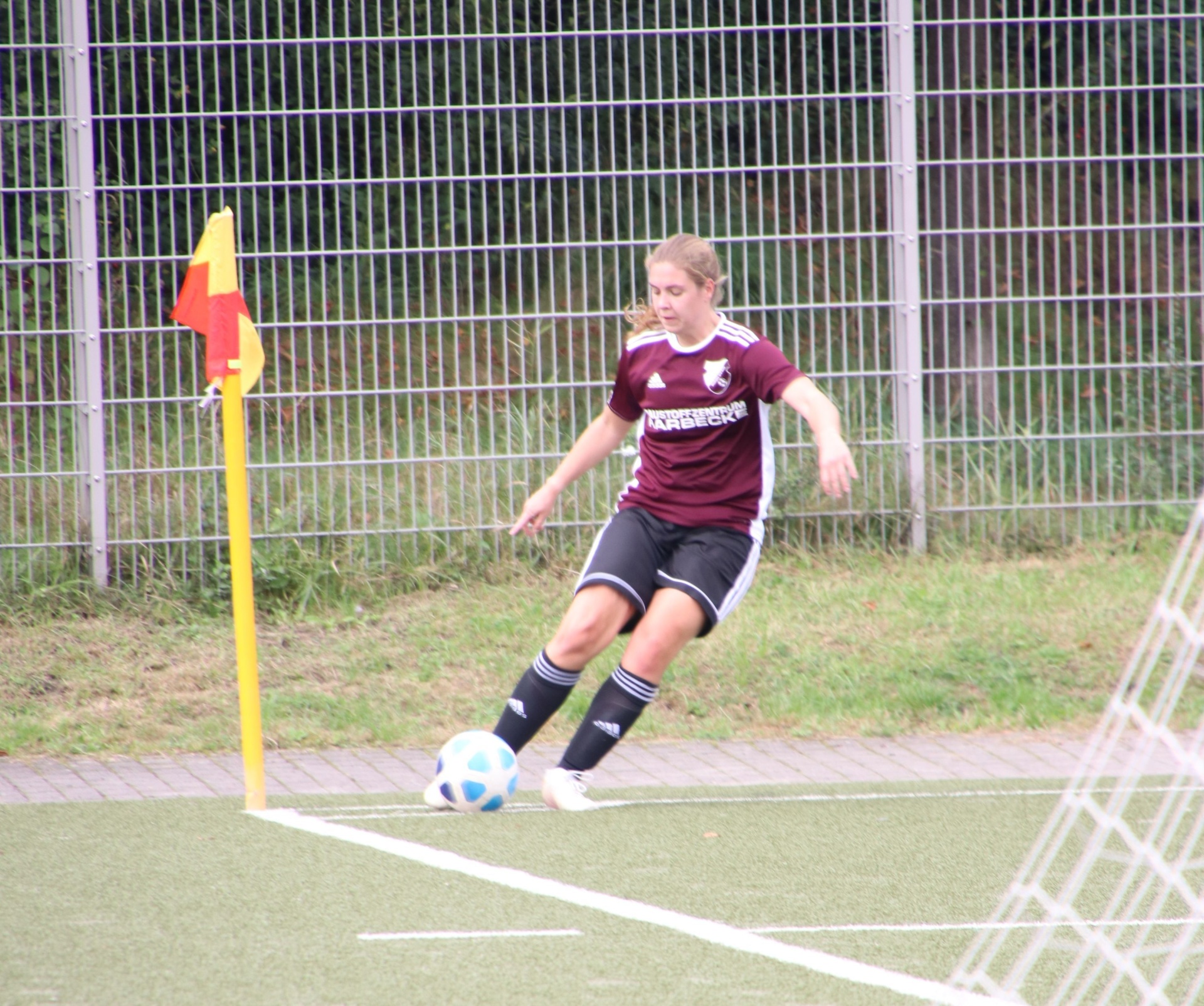 [{"clang_id":1,"value":"Fußballspielerin mit dunkelrotem Trikot und schwarzer Hose dribbelt einen Ball am Eckfeld eines Fußballplatzes. Im Hintergrund ein Metallzaun und Grün."}]
