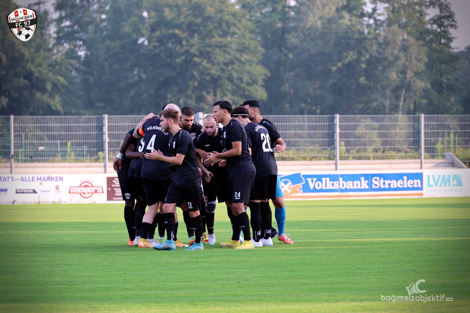[{"clang_id":1,"value":"Fußballspieler in schwarzen Trikots stehen in einem Kreis auf grünem Rasen, wahrscheinlich während einer Teambesprechung. Im Hintergrund eine Zuschauertribüne und Werbebanner."}]