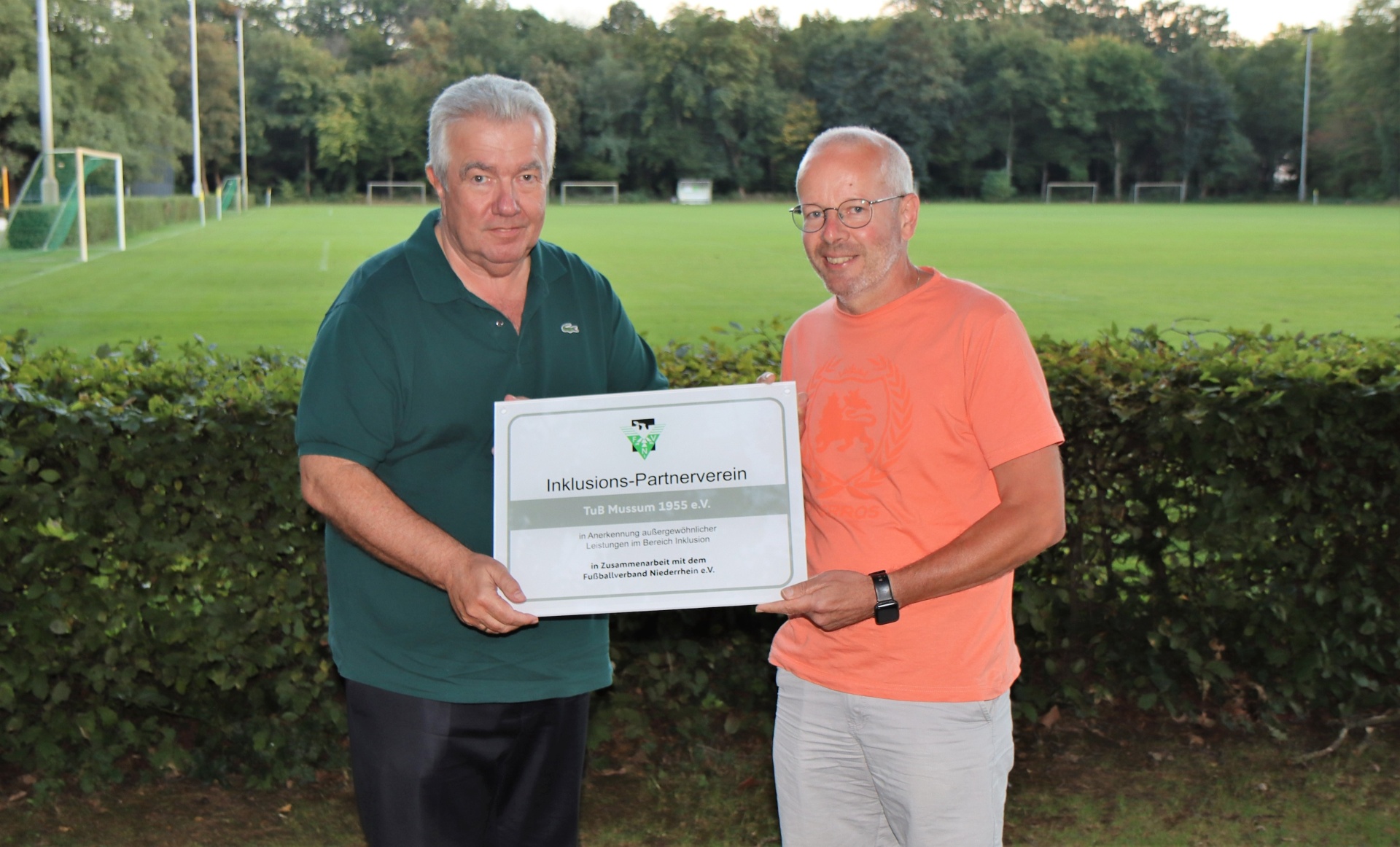 [{"clang_id":1,"value":"Zwei Männer stehen lächelnd vor einem Fußballfeld. Der linke trägt ein grünes Polohemd, der rechte ein orangefarbenes. Sie halten eine graue Plakette mit Schriftzug."}]