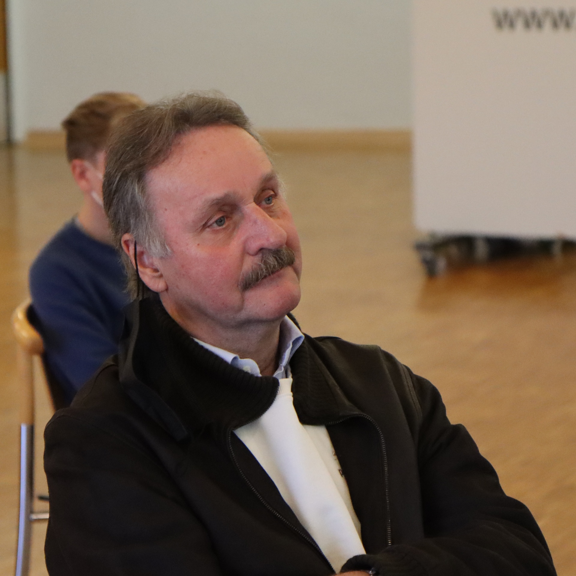 [{"clang_id":1,"value":"Mann mit grau meliertem Haar und Schnurrbart, trägt schwarze Jacke über weißem Hemd, blickt nach rechts. Im Hintergrund verschwommene Personen und ein Raum."}]