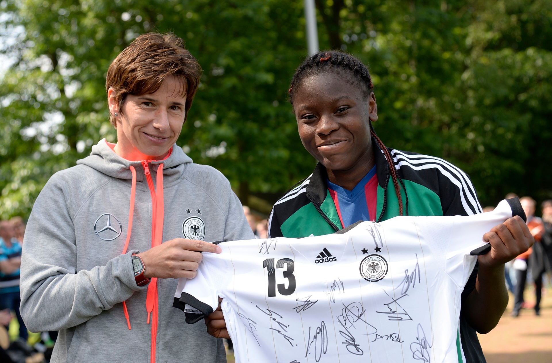 [{"clang_id":1,"value":"Zwei Frauen mit Sportbekleidung halten ein signiertes weißes Fußballtrikot mit der Nummer 13. Links eine Frau mit kurzen braunen Haaren, rechts eine Frau mit Zöpfen. Hintergrund: Grünfläche und Personen."}]