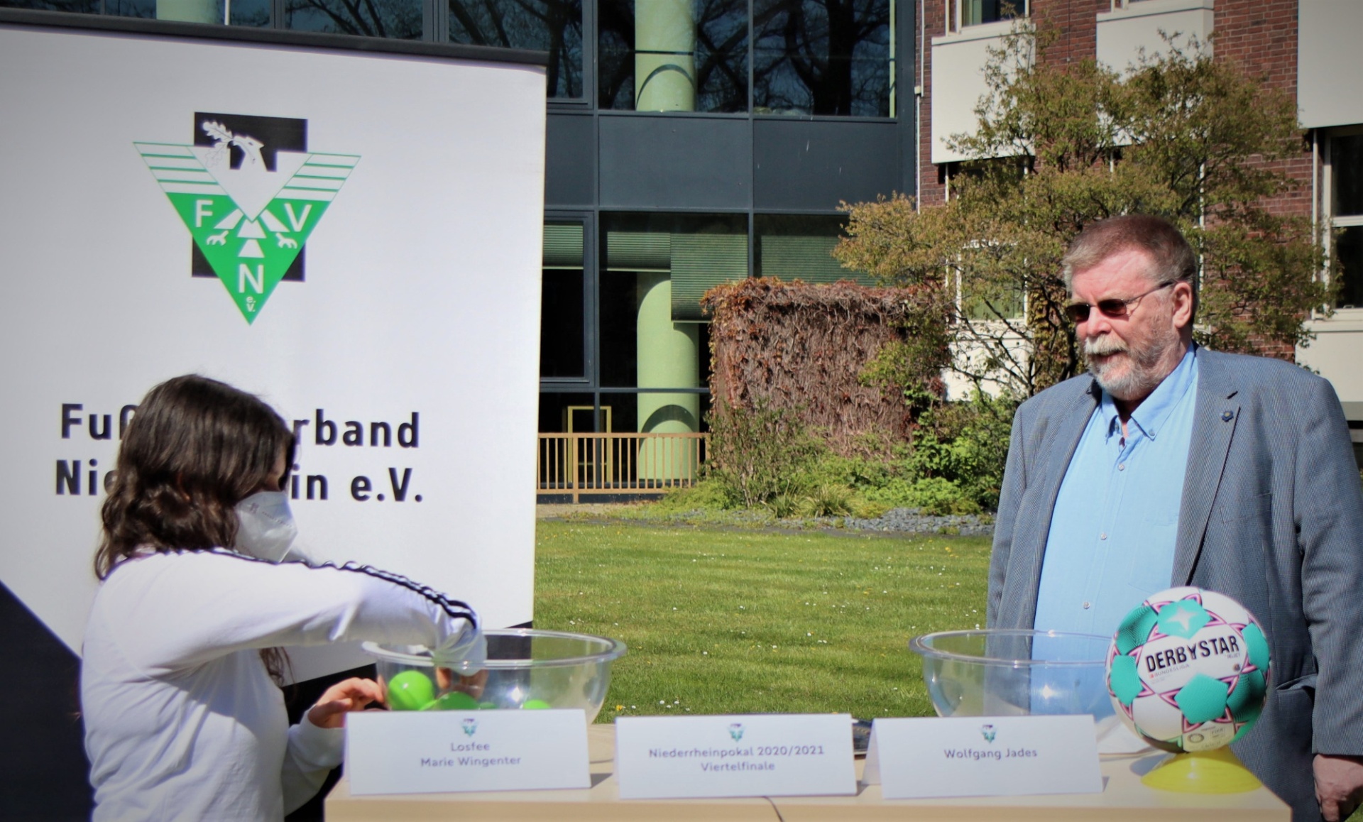 [{"clang_id":1,"value":"Zwei Personen bei einer Veranstaltung im Freien. Links ein Mädchen mit Tennisschläger und Ball, rechts ein älterer Mann mit Fußball. Im Hintergrund ein Banner und Gebäude."}]
