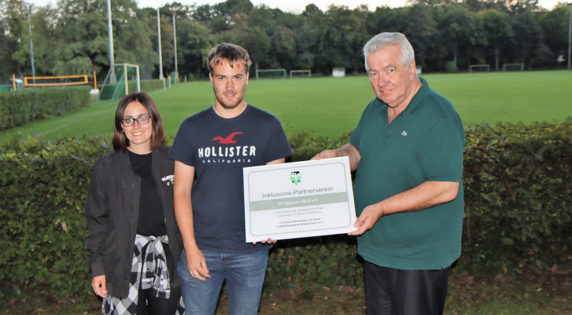 [{"clang_id":1,"value":"Drei Personen stehen vor einem Fußballfeld und halten ein Schild mit Schrift z.B. \"Inklusions-Partnerverein\" hoch. Links eine Frau, in der Mitte und rechts Männer."}]