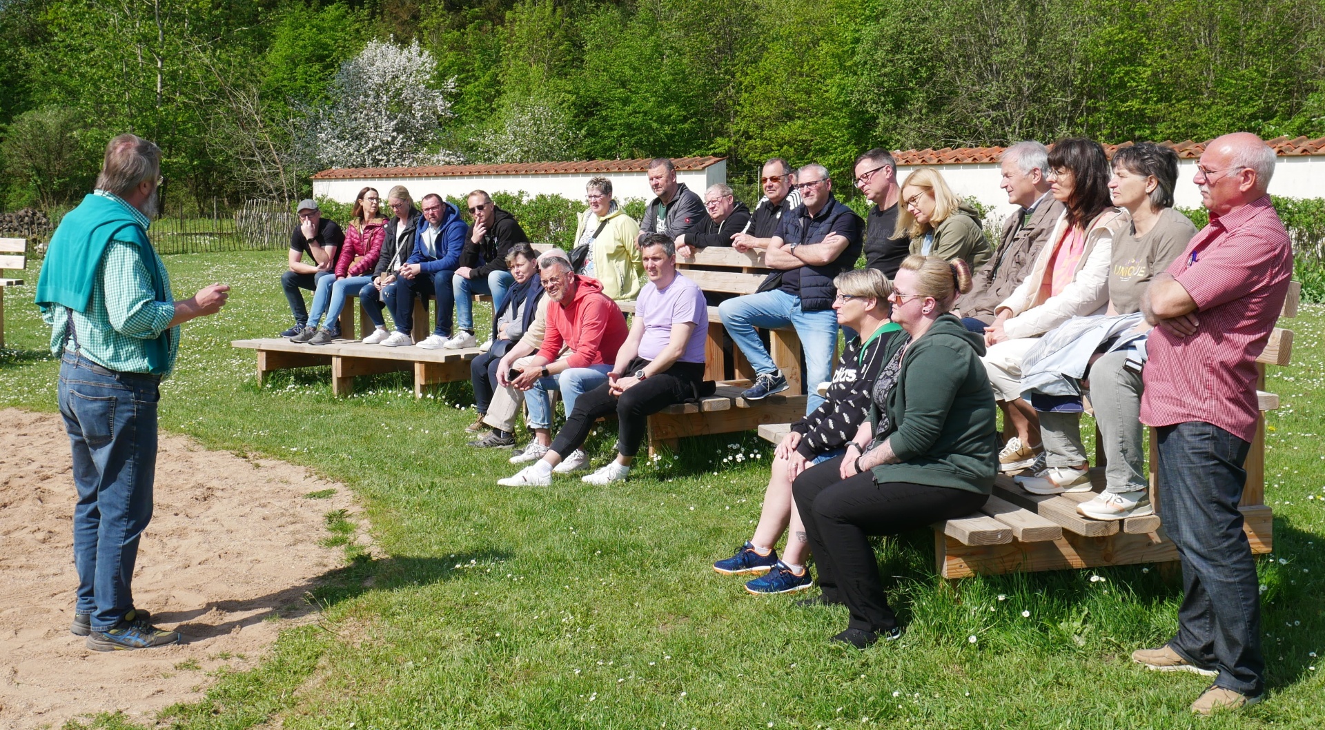[{"clang_id":1,"value":"Gruppe von etwa 20 Personen sitzt auf Bänken und steht auf einer Wiese. Ein Mann hält vor der Gruppe eine Rede. Bäume im Hintergrund."}]
