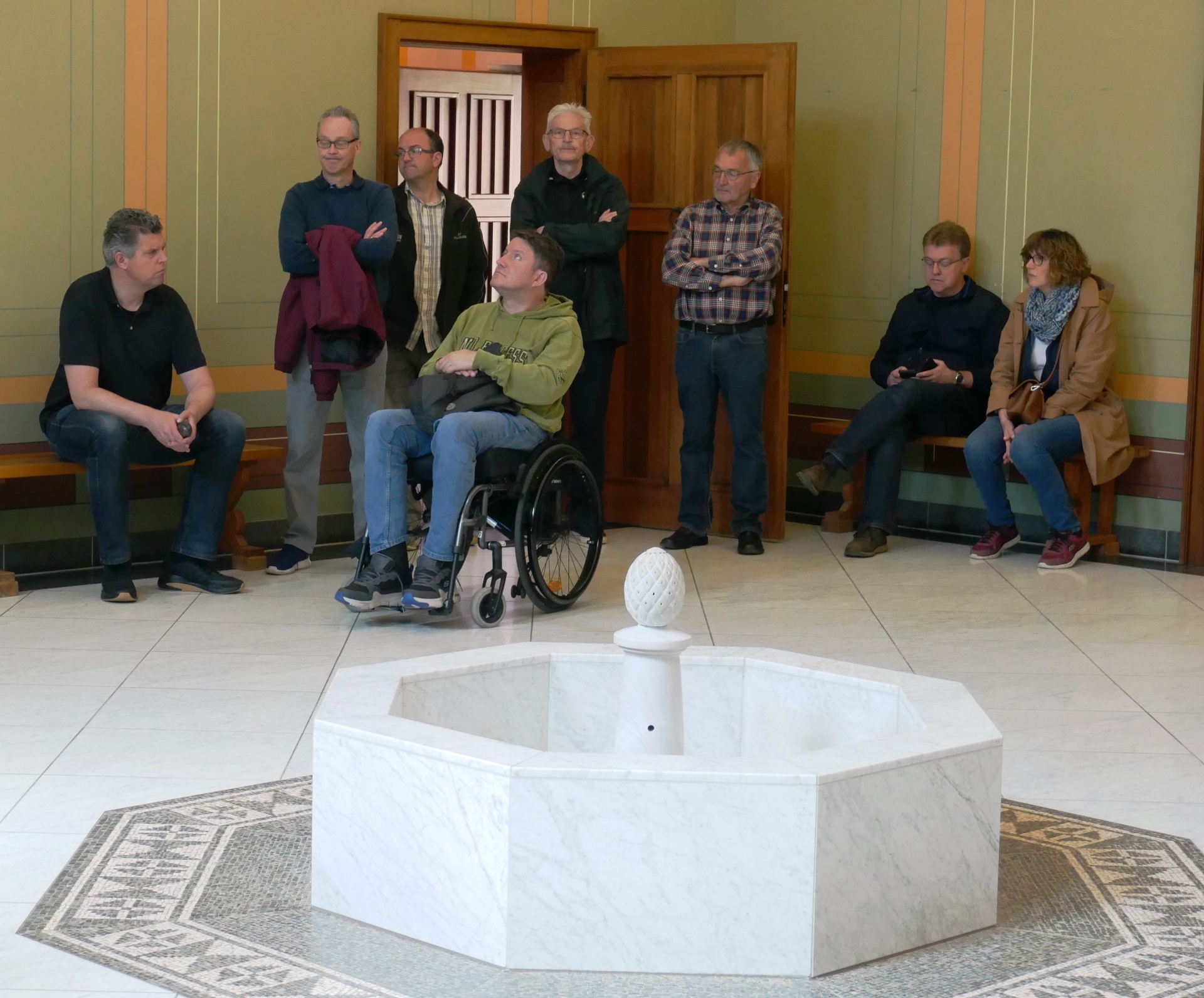 [{"clang_id":1,"value":"Gruppe Menschen in einem hellen Raum mit Marmorboden und -brunnen. Ein Mann sitzt im Rollstuhl, andere stehen oder sitzen auf Bänken."}]