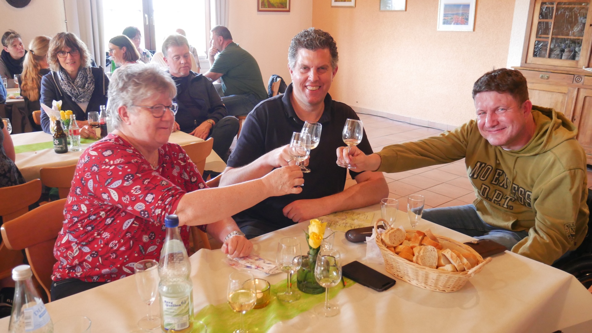 [{"clang_id":1,"value":"Drei Personen stoßen mit Weingläsern an einem gedeckten Tisch an. Im Hintergrund sind weitere Menschen in einem Restaurant zu sehen."}]