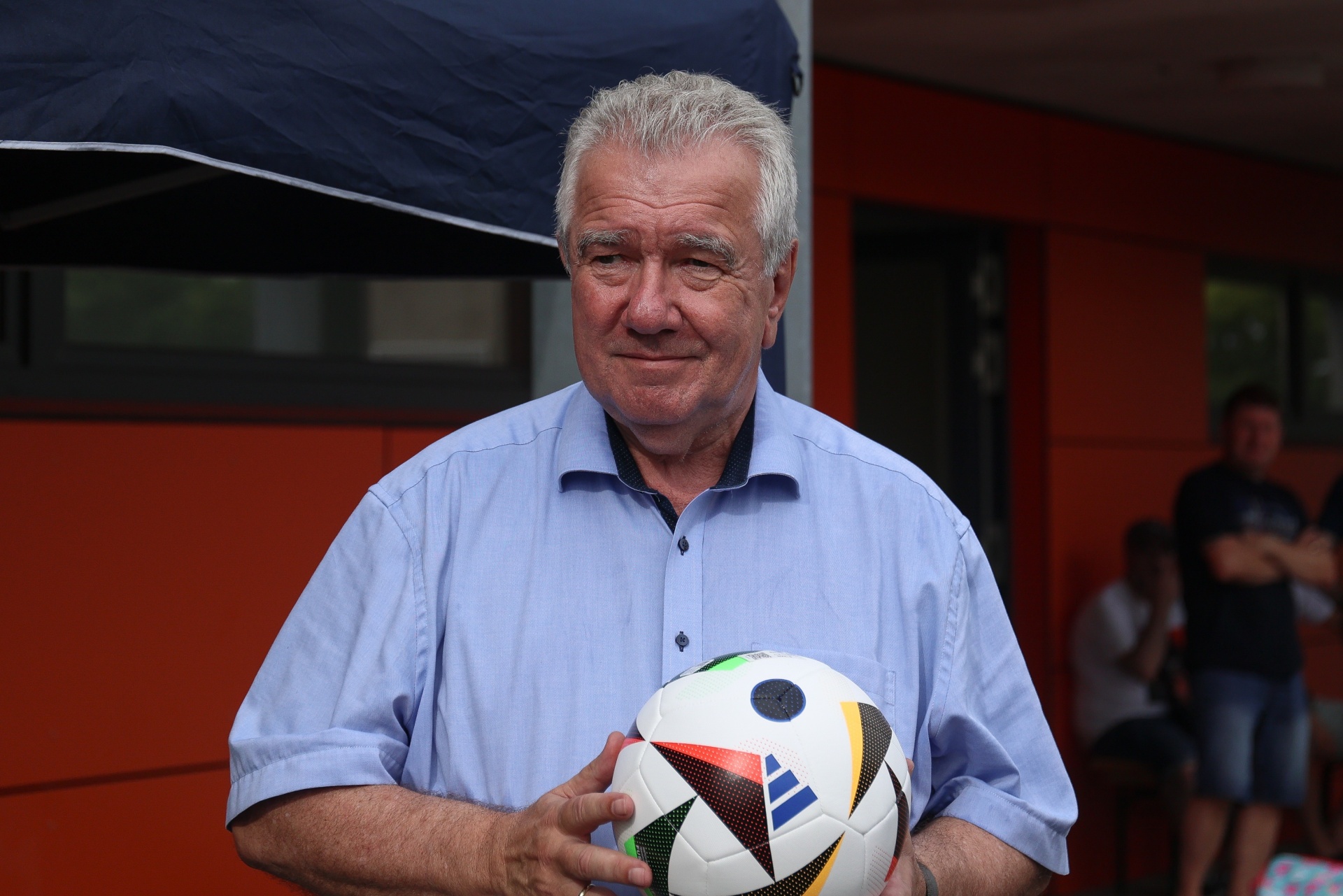 [{"clang_id":1,"value":"Älterer Mann mit grauem Haar hält einen Fußball in den Händen. Er trägt ein hellblaues Hemd. Im Hintergrund Personen und ein roter Hintergrund."}]