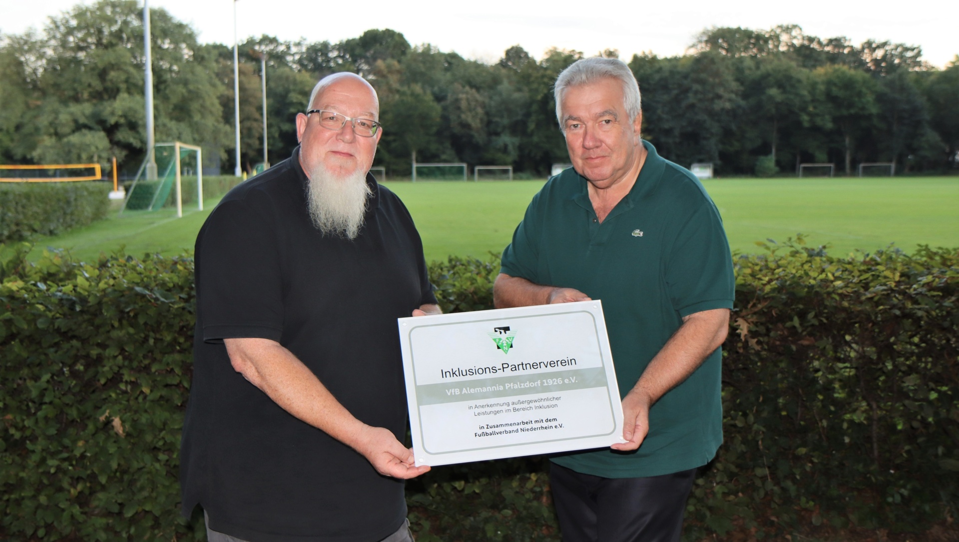[{"clang_id":1,"value":"Zwei Männer stehen auf einer Grünfläche vor einem Fußballfeld. Der linke Mann hat einen Bart und trägt Schwarz, der rechte einen grünen Pullover und hält eine Plakette mit Aufschrift."}]