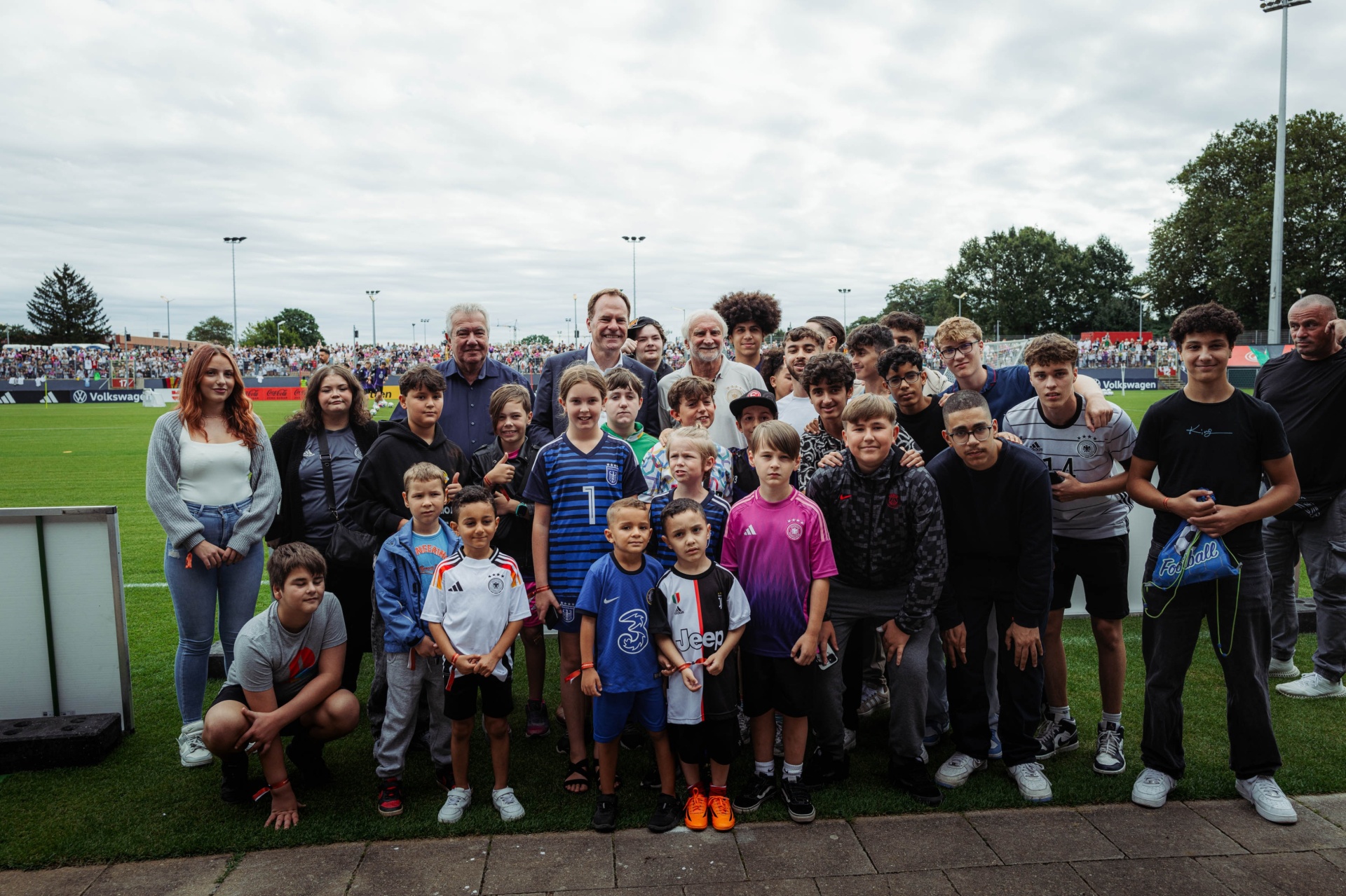 [{"clang_id":1,"value":"Gruppe mit etwa 30 Personen, darunter viele Kinder und Jugendliche, vor Fußballstadion bei bewölktem Himmel. Einige tragen Fußballtrikots."}]