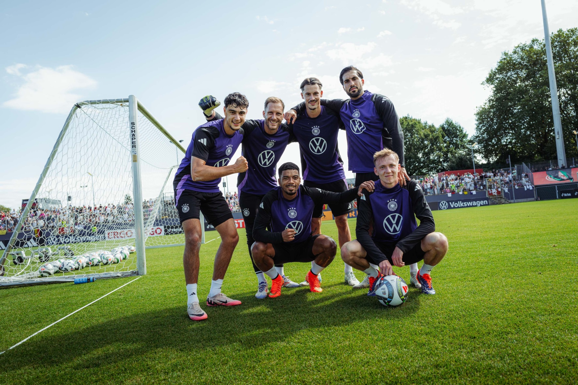 [{"clang_id":1,"value":"Fußballmannschaft in violetten Trainingsanzügen posiert lächelnd auf dem Rasen vor einem Fußballtor und Zuschauerrängen. Volkswagen-Werbung im Hintergrund."}]