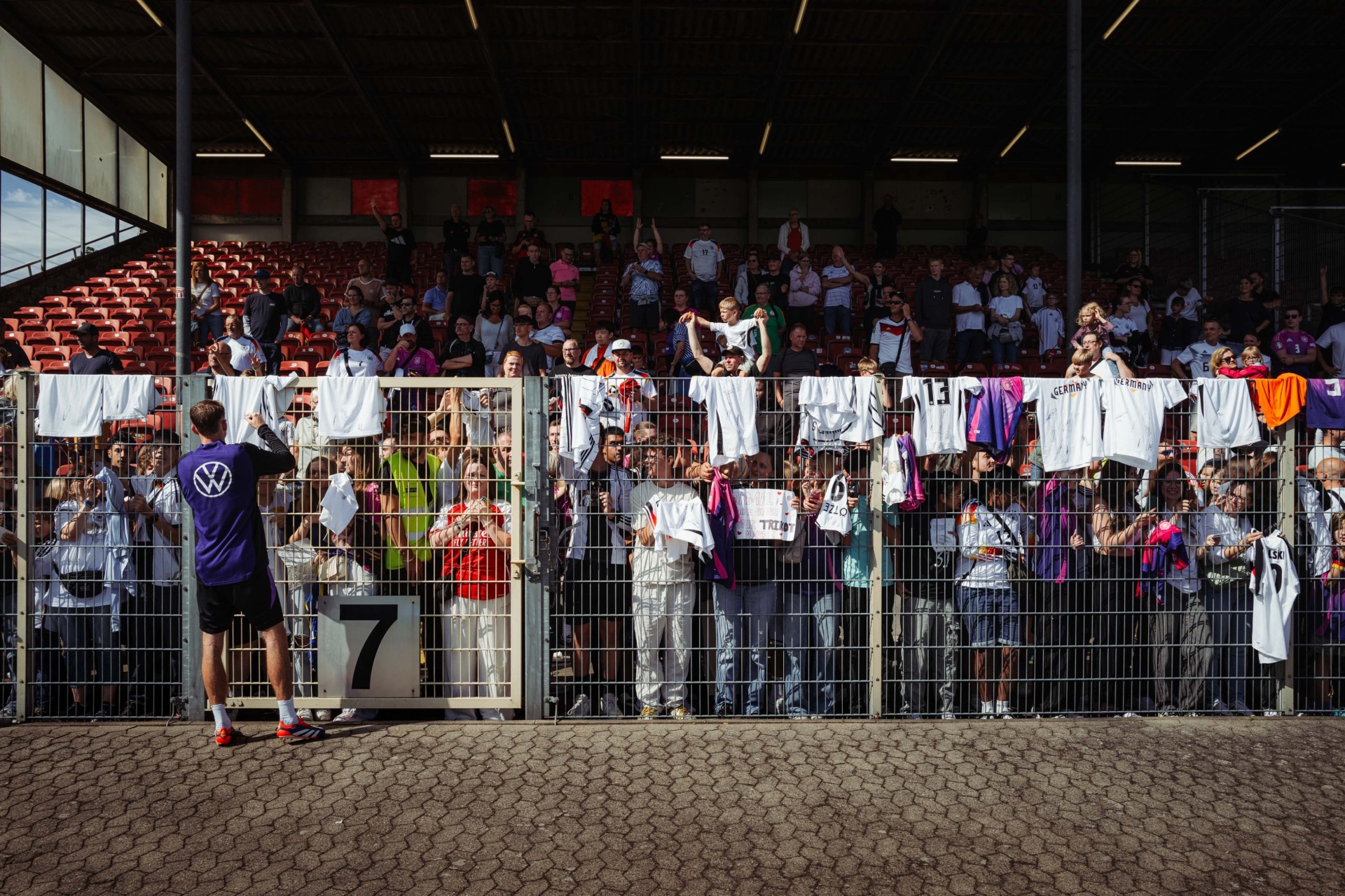 [{"clang_id":1,"value":"Fußballstadion: Spieler mit Startnummer 7 steht vor Zuschauermenge an einem Zaun. Menschen sitzen auf roten und orangen Tribünen."}]