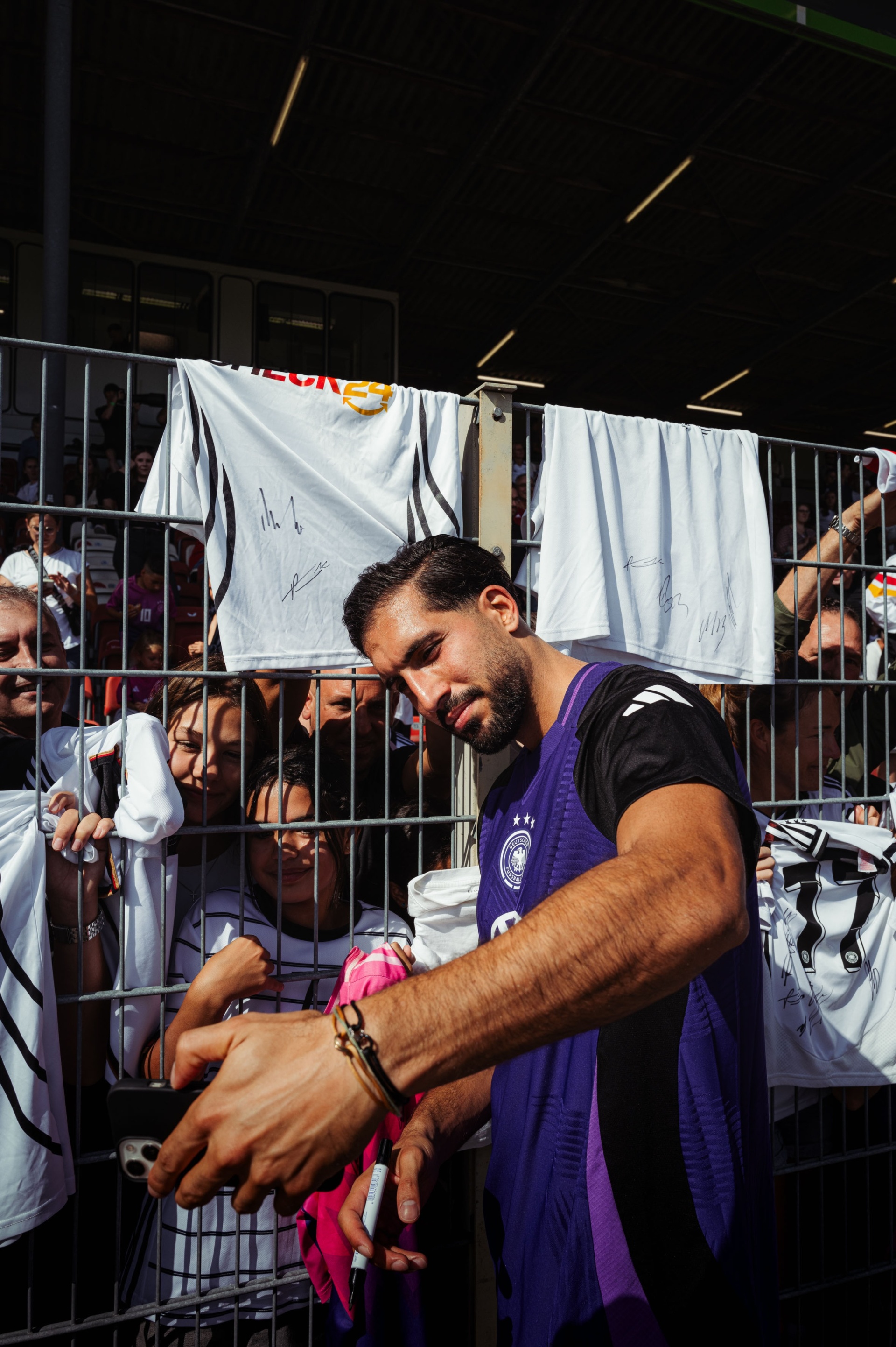 [{"clang_id":1,"value":"Fußballspieler mit Bart unterschreibt Trikots und Autogrammwünsche an Fans hinter einer Absperrung. Hintergrund ist eine Menschenmenge und ein Stadion."}]