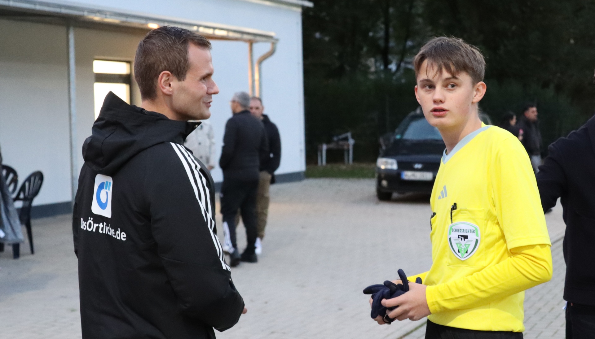 [{"clang_id":1,"value":"Zwei Männer stehen draußen vor einem Gebäude. Links ein Mann mit schwarzer Sportjacke, rechts ein Jugendlicher im Fußballtrikot. Im Hintergrund weitere Personen und Autos."}]