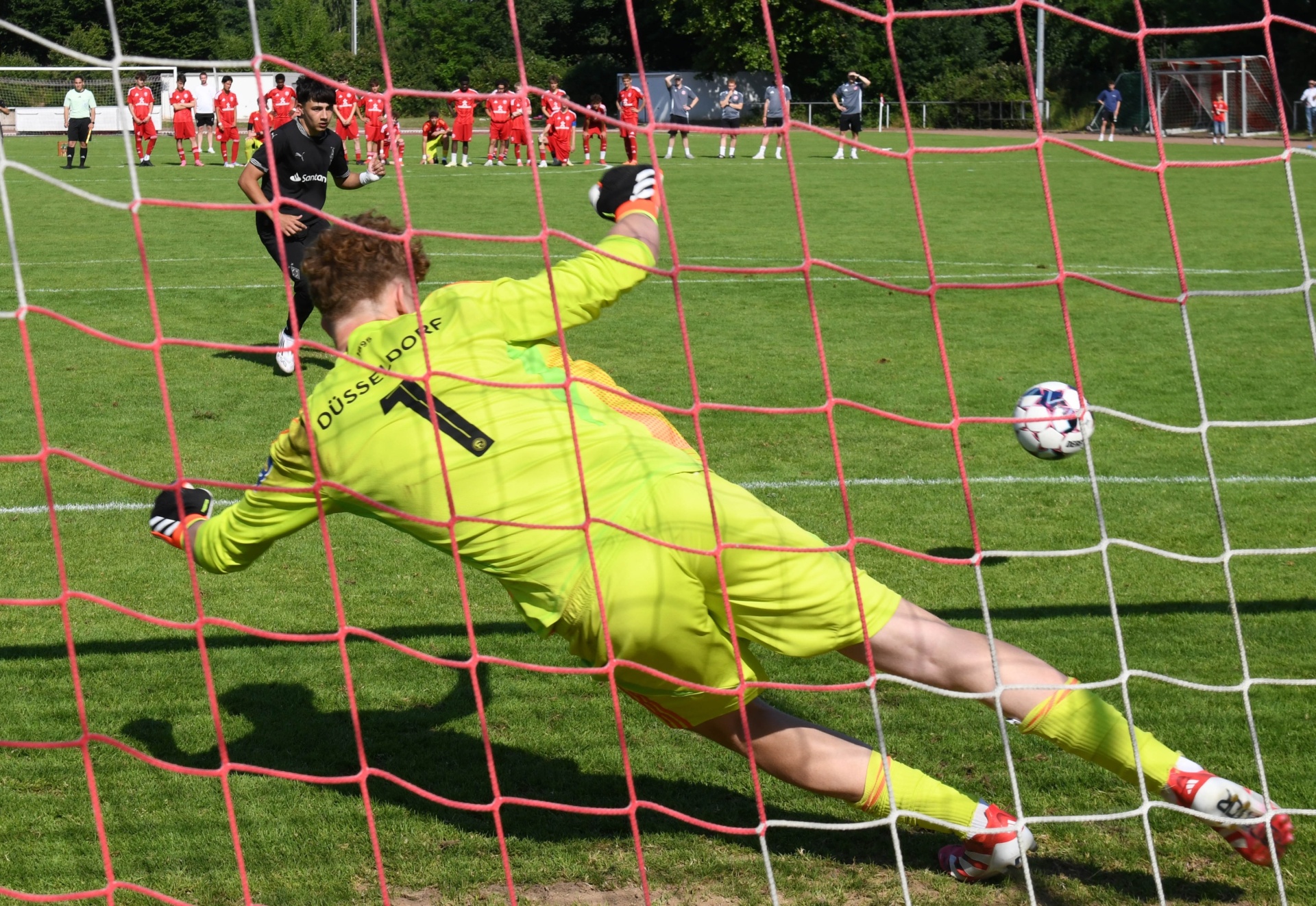 [{"clang_id":1,"value":"Fußballtorwart in gelbem Trikot springt nach Ball, der sich im Netz befindet. Im Hintergrund stehen Fußballspieler auf einem grünen Rasen."}]