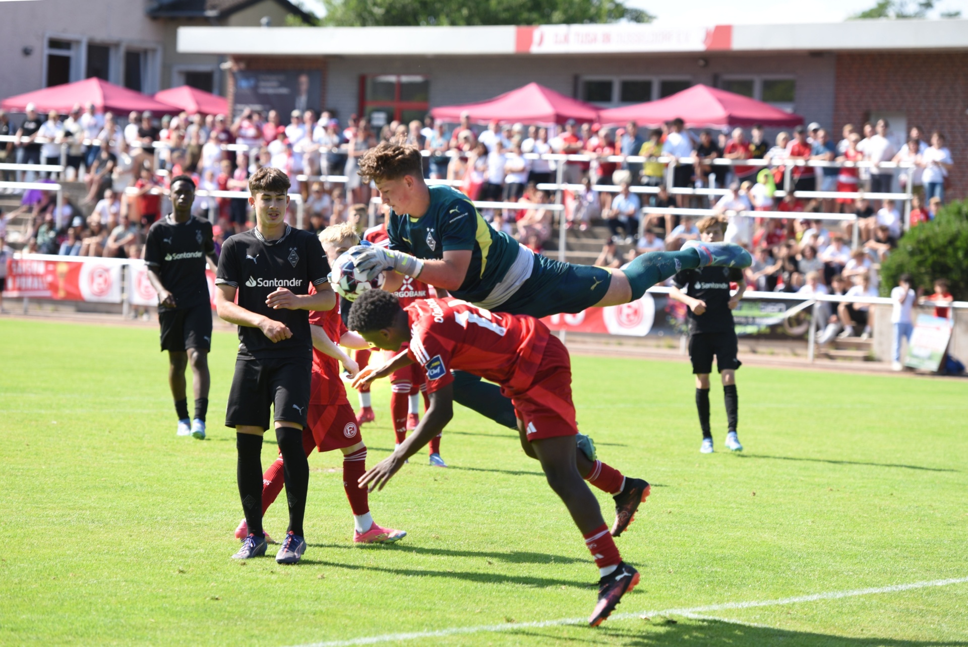 [{"clang_id":1,"value":"Fußballspiel: Torwart springt nach rechts, um Ball zu fangen. Zwei Spieler in Rot und Schwarz stehen davor, Hintergrund mit Zuschauertribüne."}]