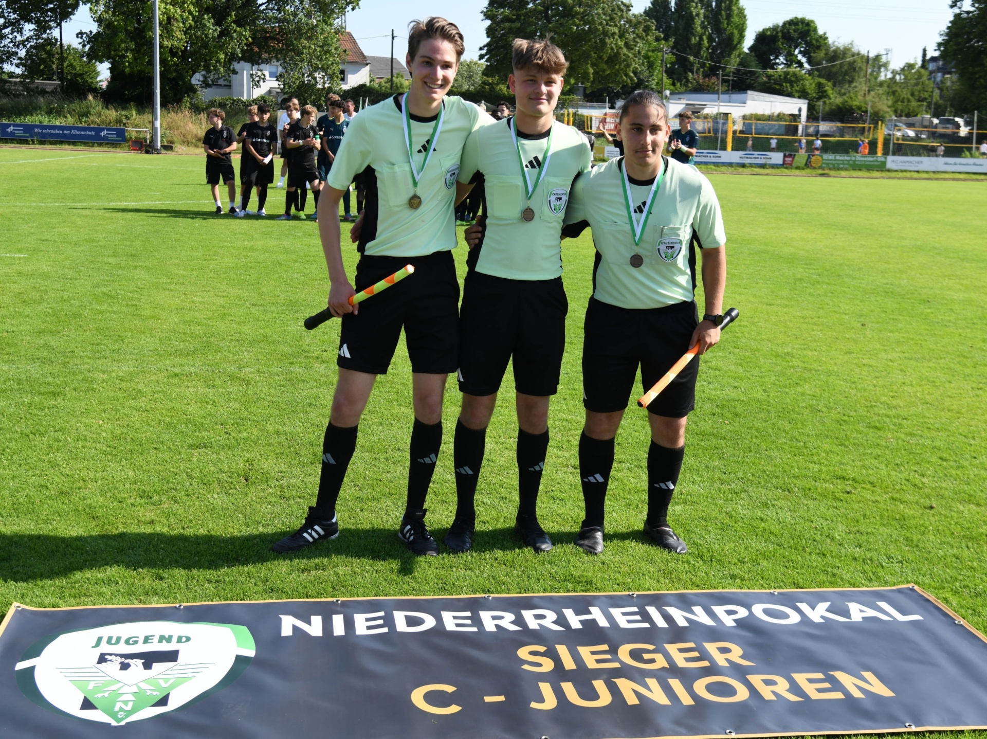 [{"clang_id":1,"value":"Drei junge Bandyspieler mit Medaillen und Schlägern stehen auf einem grünen Rasen vor einem schwarzen Banner mit weißer Schrift: \"NIEDERRHEINPOKAL SIEGER C-JUNIOREN\". Im Hintergrund weitere Sportler und Gebäude."}]