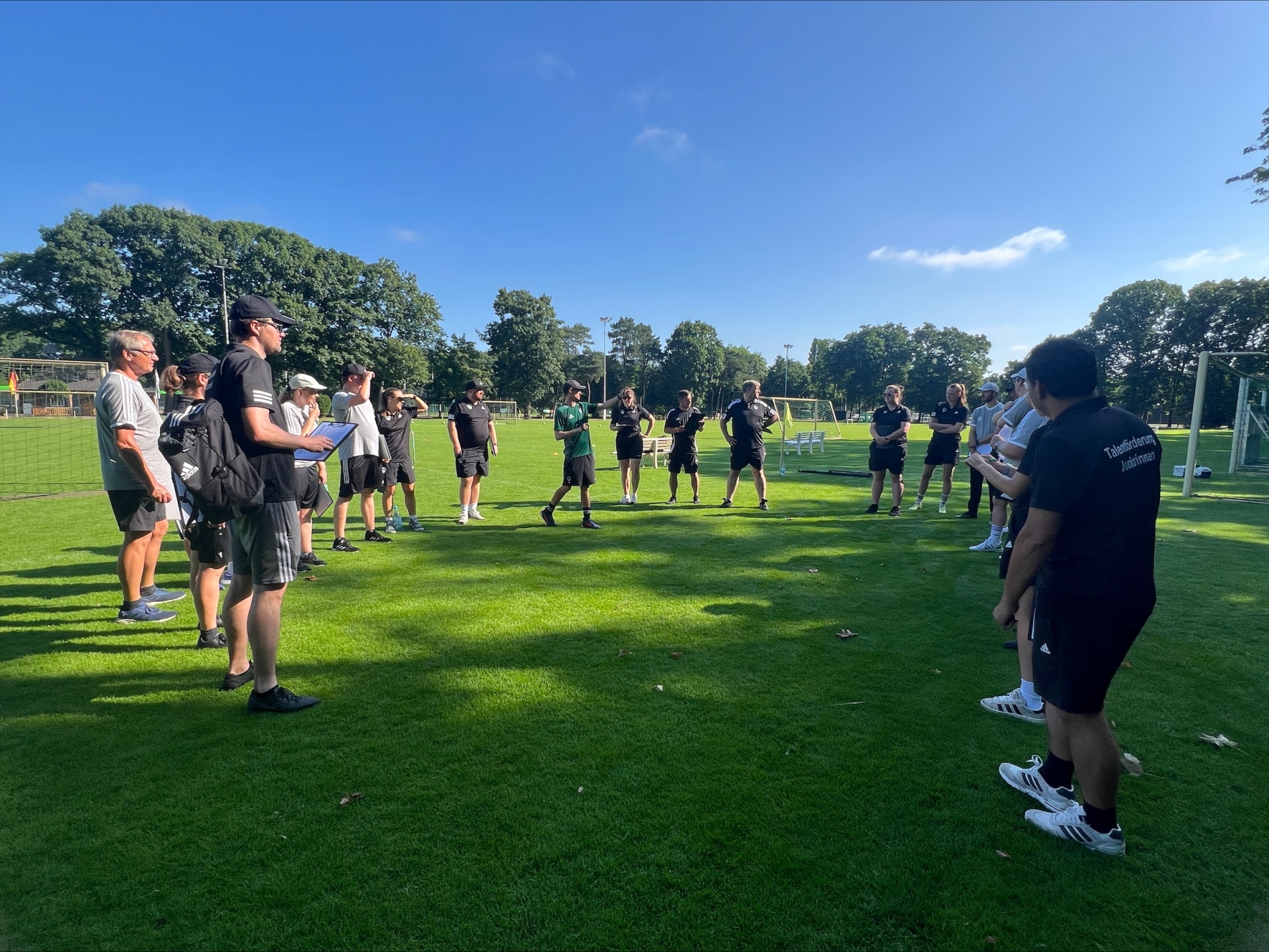 [{"clang_id":1,"value":"Fußballteam im Kreis auf grünem Rasen. Trainer gibt Anweisungen. Hintergrund: Bäume und blauer Himmel."}]