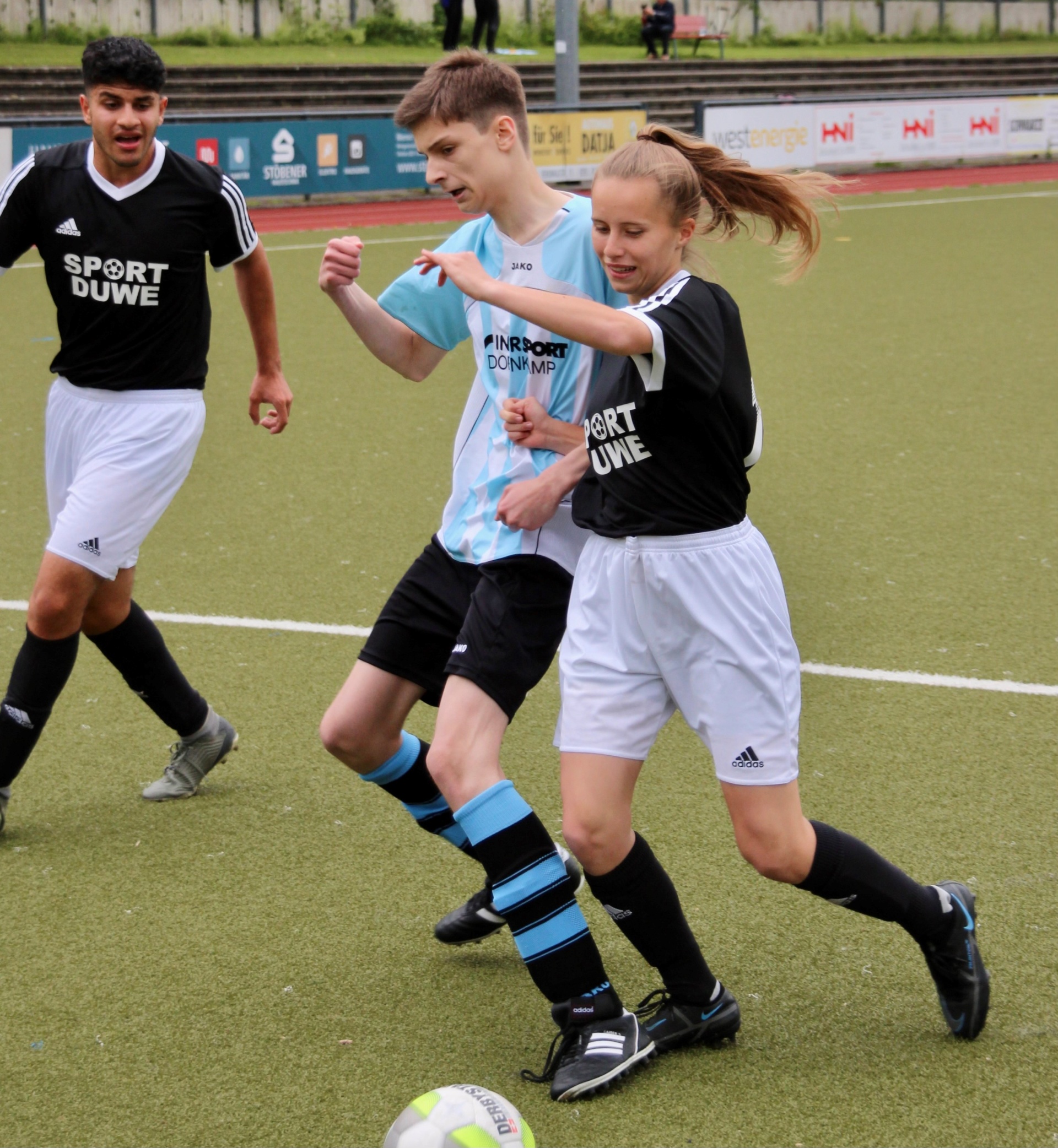 [{"clang_id":1,"value":"Drei Jugendliche spielen Fußball auf einem Rasenplatz. Ein Mädchen in schwarzem Trikot und Shorts dribbelt den Ball, während zwei Jungen sie stellen."}]