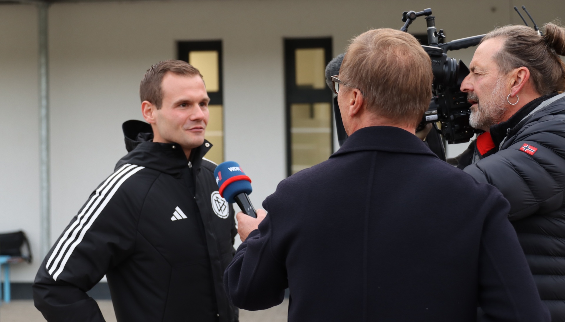 [{"clang_id":1,"value":"Mann in Sportjacke wird interviewt. Kamerateam filmt die Szene vor weißem Hintergrund. Ein Mann trägt eine rote Jacke mit dänischer Flagge."}]