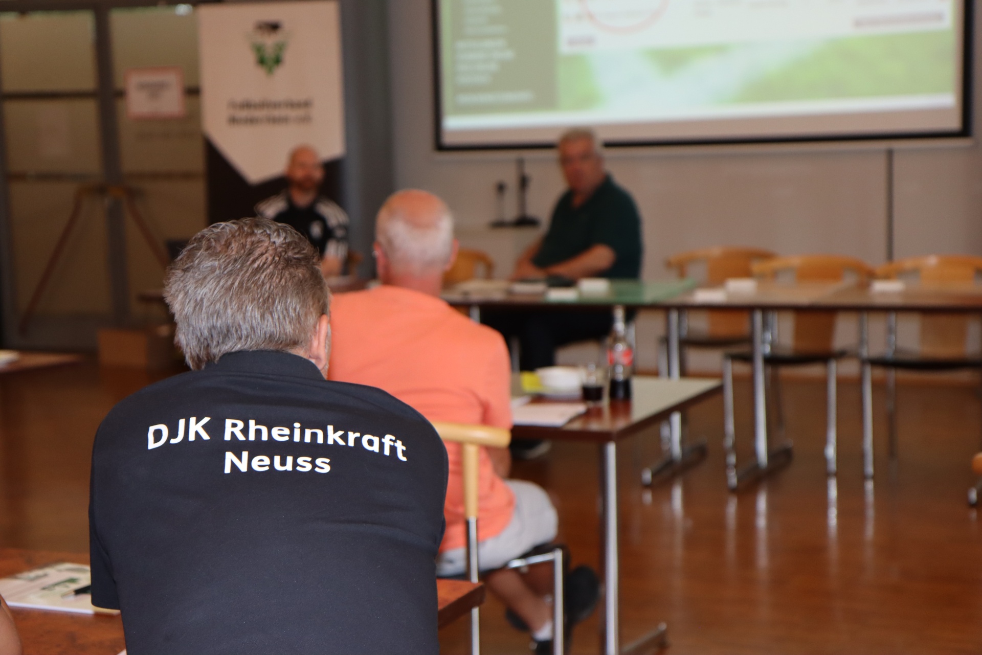 [{"clang_id":1,"value":"Sitzung in Turnhalle. Im Vordergrund Mann mit T-Shirt \"DJK Rheinkraft Neuss\" mit dem Rücken zum Betrachter. Im Hintergrund Menschen an Tischen, Projektion an Wand."}]
