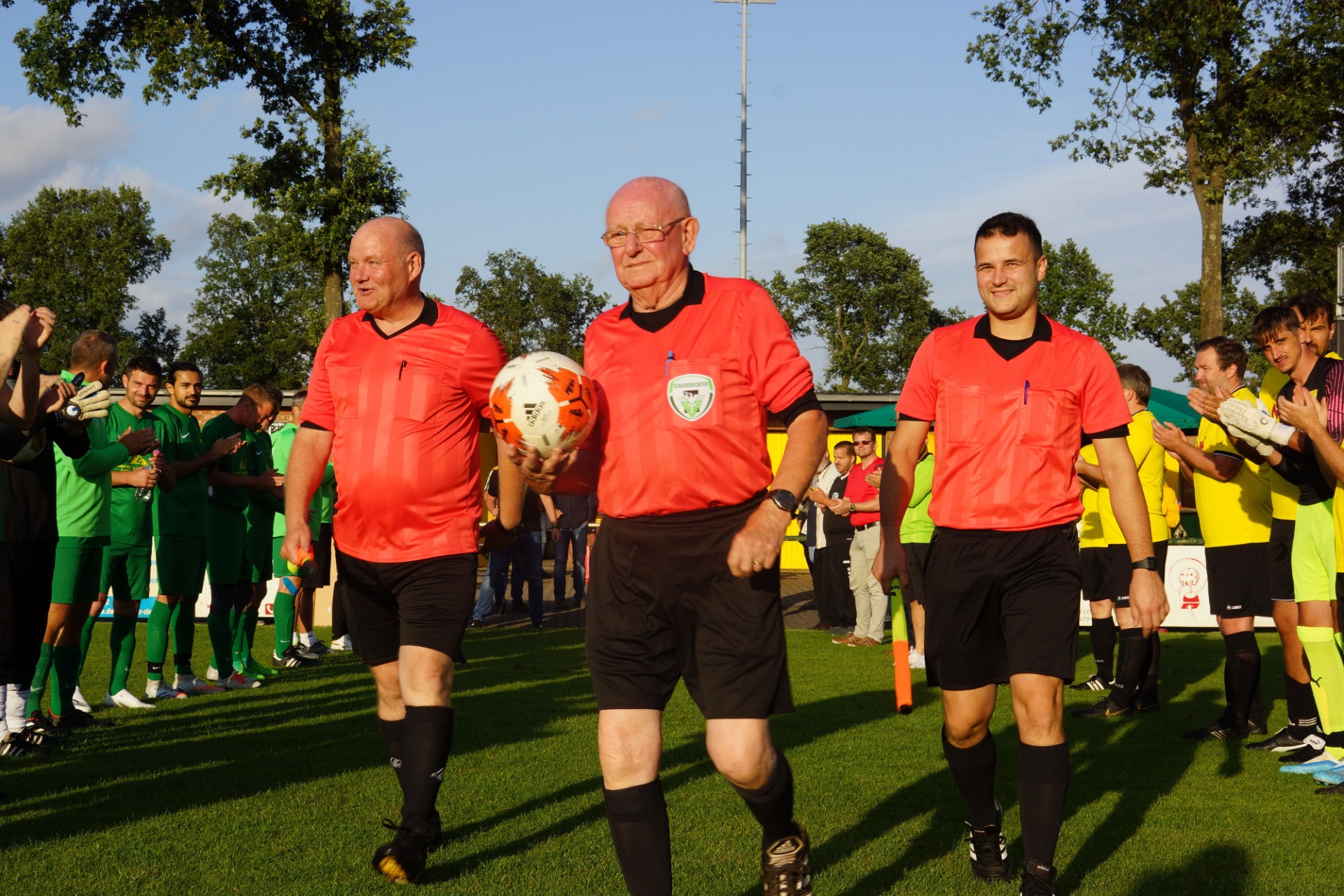 [{"clang_id":1,"value":"Drei Schiedsrichter in roten Trikots und schwarzen Shorts laufen auf einem Fußballfeld. Im Hintergrund stehen Spieler in grünen und gelben Trikots."}]