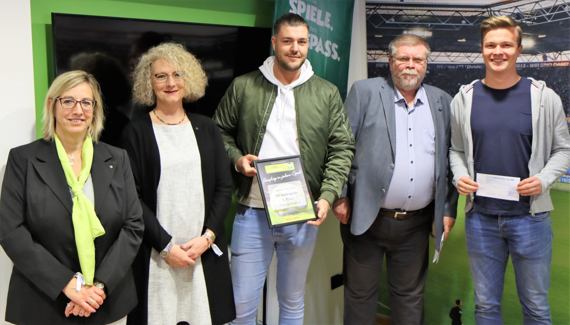 [{"clang_id":1,"value":"Sechs Personen stehen nebeneinander in einem Innenraum. Drei Frauen und drei Männer halten Urkunden oder Auszeichnungen. Im Hintergrund eine grüne Wand mit Text und eine Stadionkulisse."}]