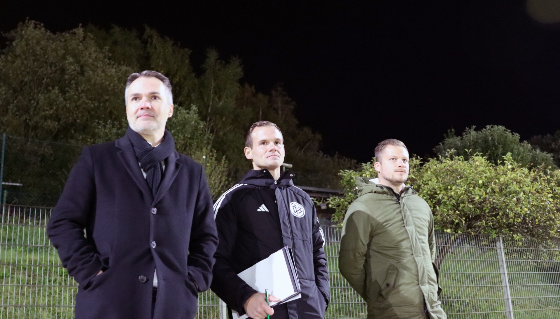 [{"clang_id":1,"value":"Drei Männer stehen nebeneinander auf einem Sportplatz im Dunkeln. Links trägt einer einen schwarzen Mantel, in der Mitte einen schwarzen Trainingsanzug und rechts eine grüne Jacke."}]