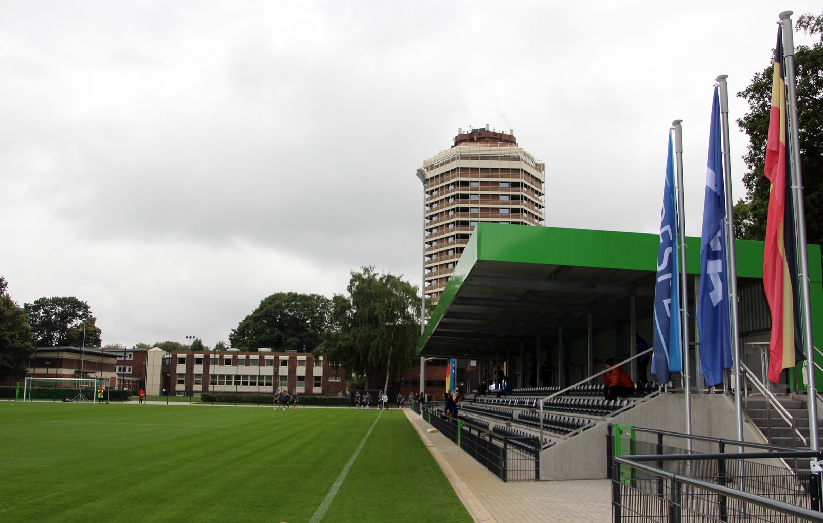 [{"clang_id":1,"value":"Fußballstadion mit grüner Rasenfläche, Tribüne mit Sitzplätzen und dahinter ein Hochhaus sowie weitere Gebäude und Bäume unter grauem Himmel."}]