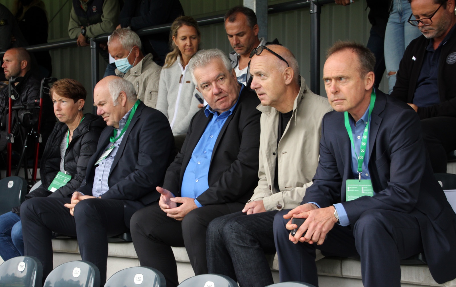 [{"clang_id":1,"value":"Gruppe von Männern und Frauen sitzt auf Stadiontribünen, viele tragen Anzüge und Lanyards. Im Hintergrund weitere Zuschauer und Stadionelemente."}]