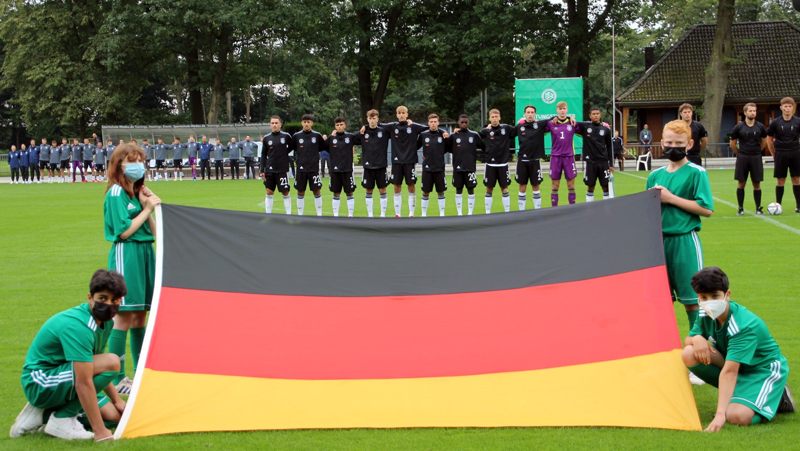 [{"clang_id":1,"value":"Fußballmannschaft in schwarzen Trikots steht mit der deutschen Flagge (Schwarz, Rot, Gold) vor einem Fußballfeld. Zuschauer im Hintergrund."}]