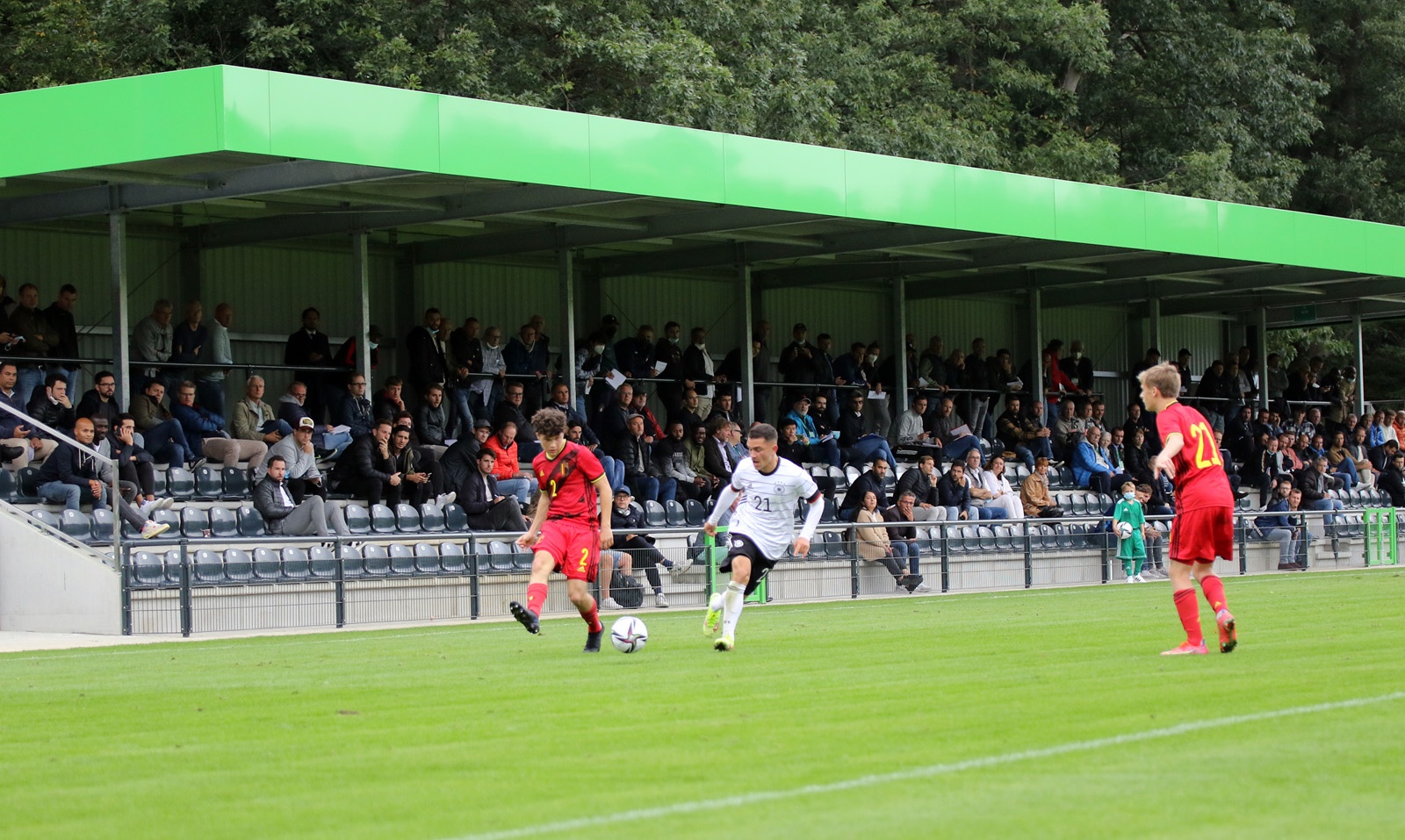 [{"clang_id":1,"value":"Fußballspiel. Drei Spieler auf grünem Rasen, zwei in rot und einer in weiß. Im Hintergrund eine Zuschauertribüne mit grünem Dach."}]