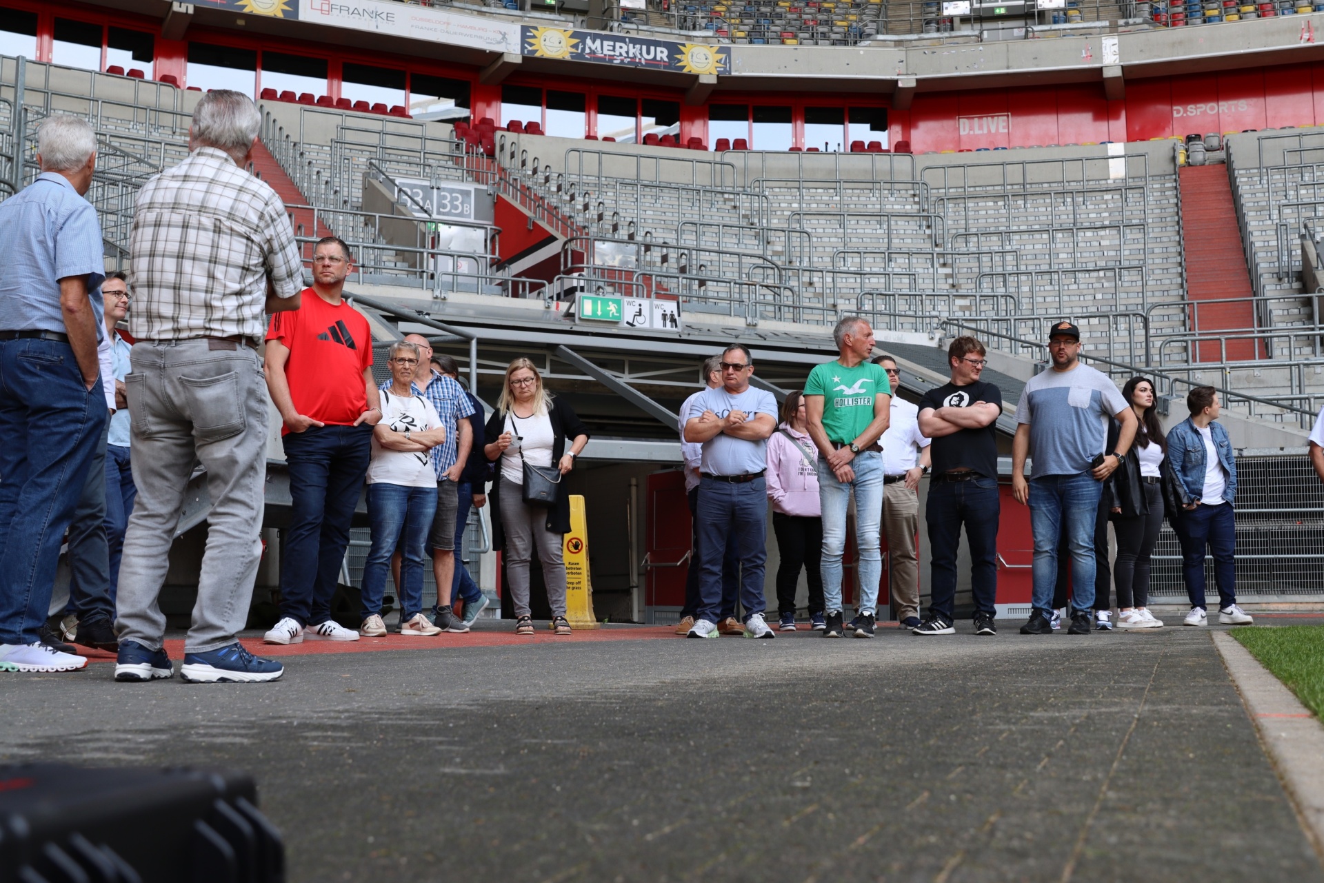 [{"clang_id":1,"value":"Gruppe Menschen steht auf einer Tribüne vor leeren Stadionrängen. Ein Mann mit kariertem Hemd spricht die Gruppe an. Im Hintergrund Werbebanner."}]