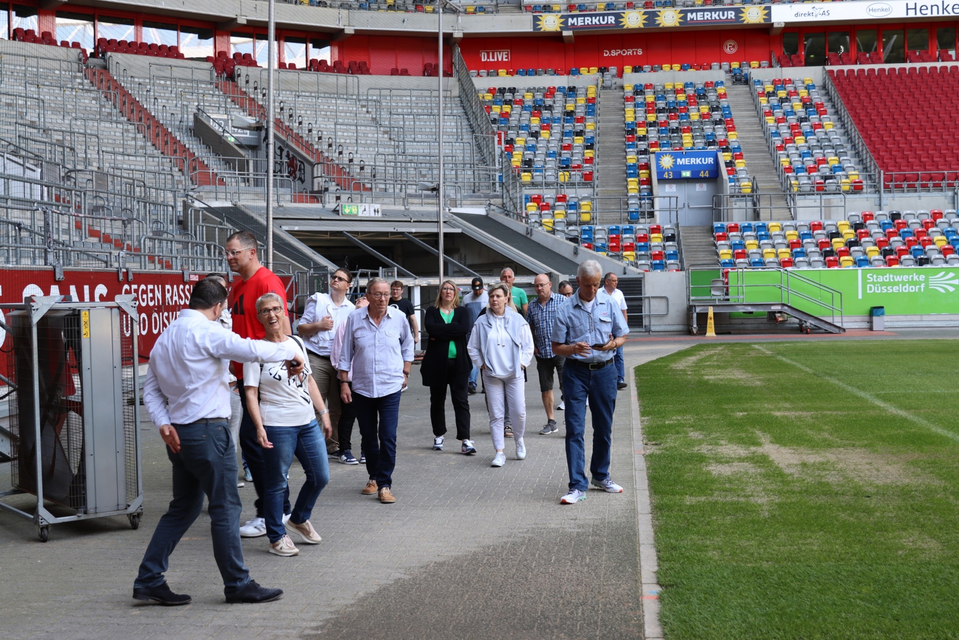 [{"clang_id":1,"value":"Fußballstadion mit Zuschauerrängen. Eine Gruppe Personen steht auf einem Gang vor dem Spielfeld und unterhält sich."}]