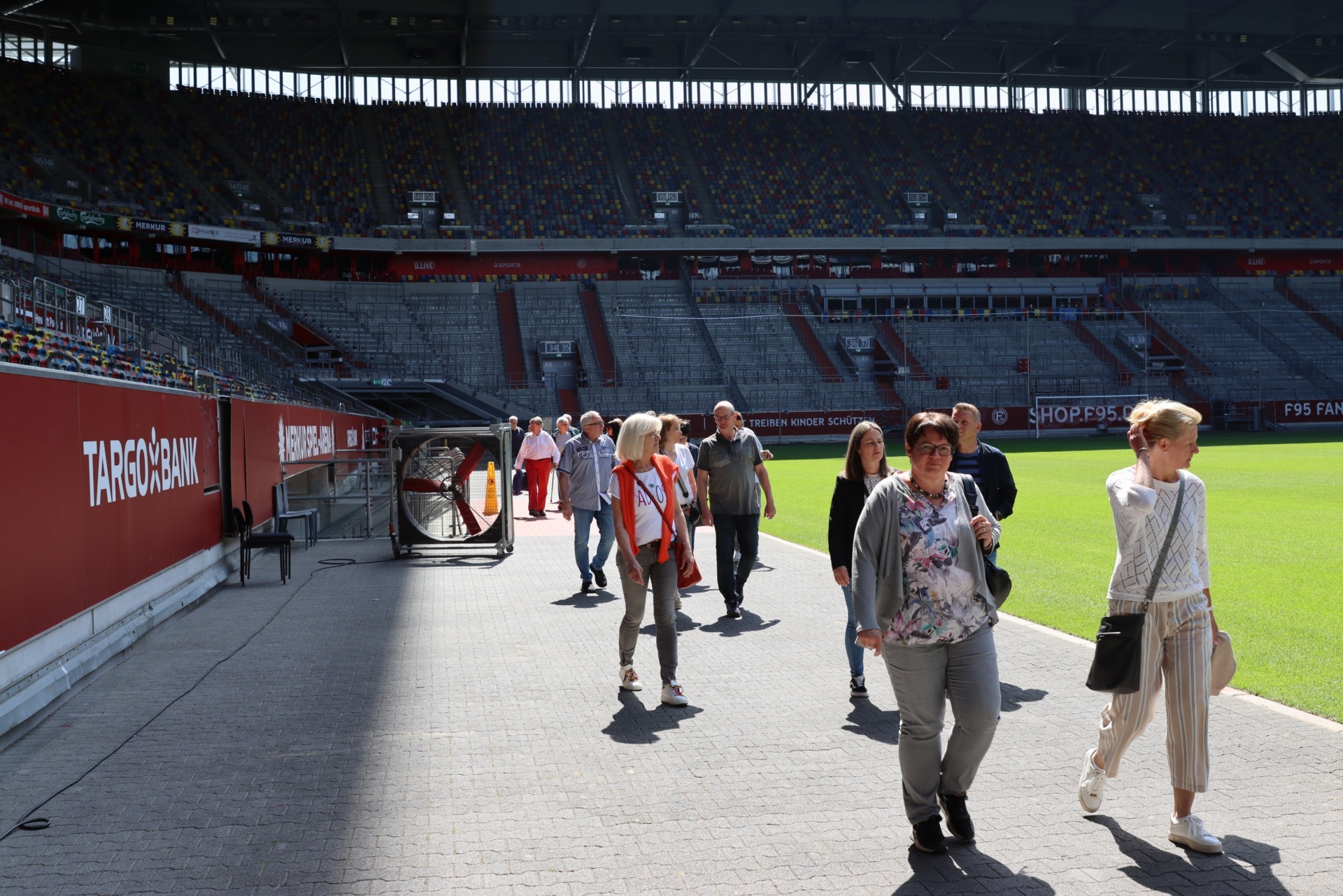 [{"clang_id":1,"value":"Fußballstadion: Menschen gehen auf einem Gehweg an den Sitztribünen vorbei zum Rasen. Im Hintergrund Werbebanner."}]