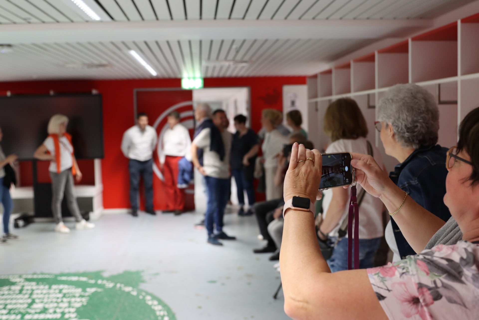 [{"clang_id":1,"value":"Personen stehen in einem Raum mit roter Wand und filmen/fotografieren eine Gruppe Männer in heller Kleidung. Im Vordergrund eine Frau mit Smartphone."}]
