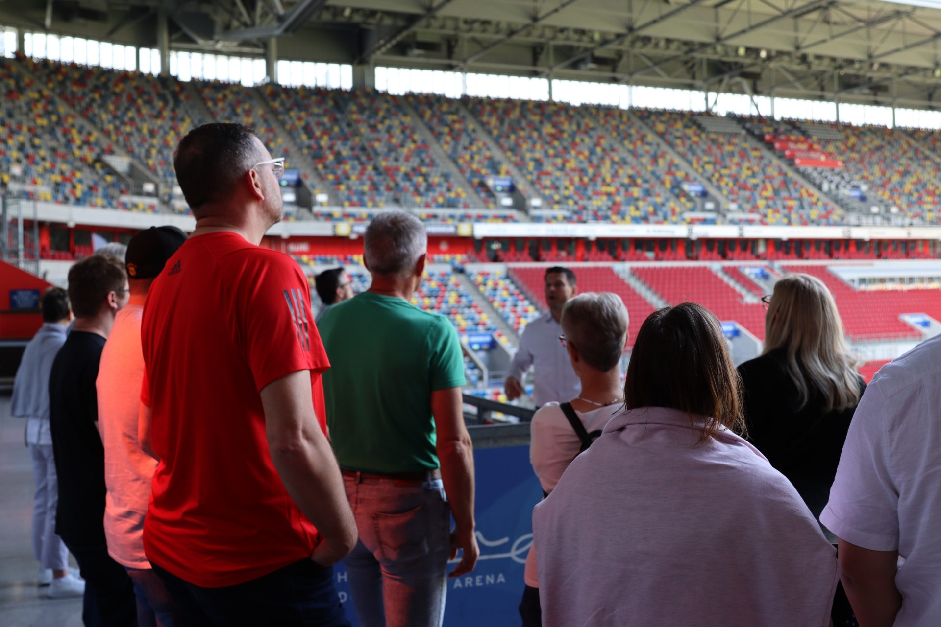[{"clang_id":1,"value":"Personengruppe steht im Stadion und blickt auf die Zuschauerränge. Im Hintergrund leere Sitze in verschiedenen Farben."}]